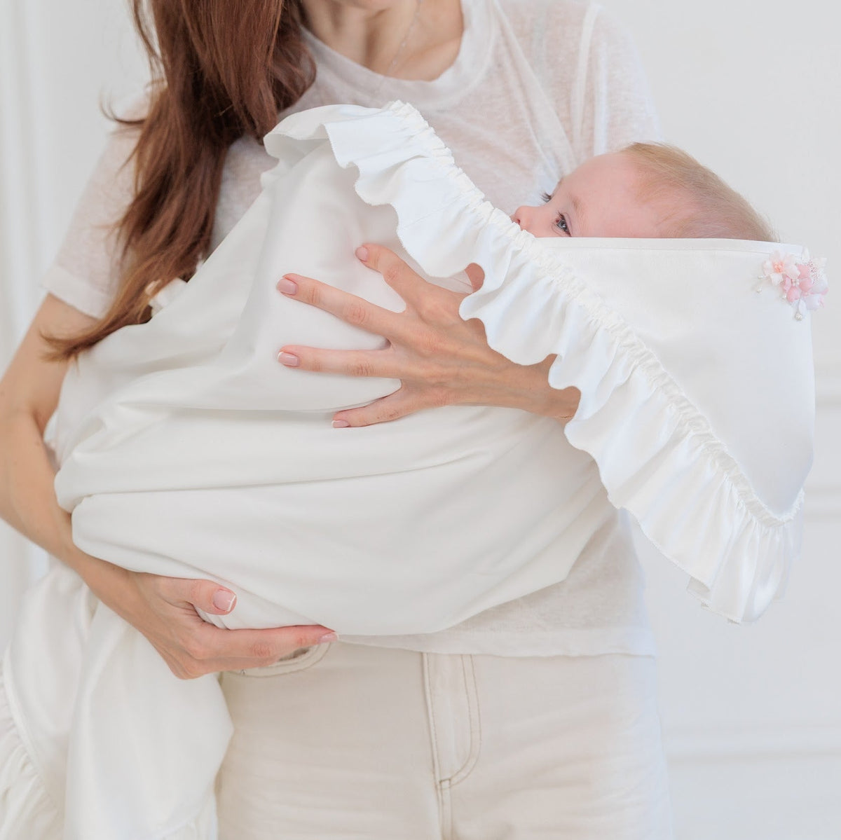 A woman cradles a baby wrapped in a soft white ruffled swaddle blanket, set against a bright, minimalist white paneled background.
