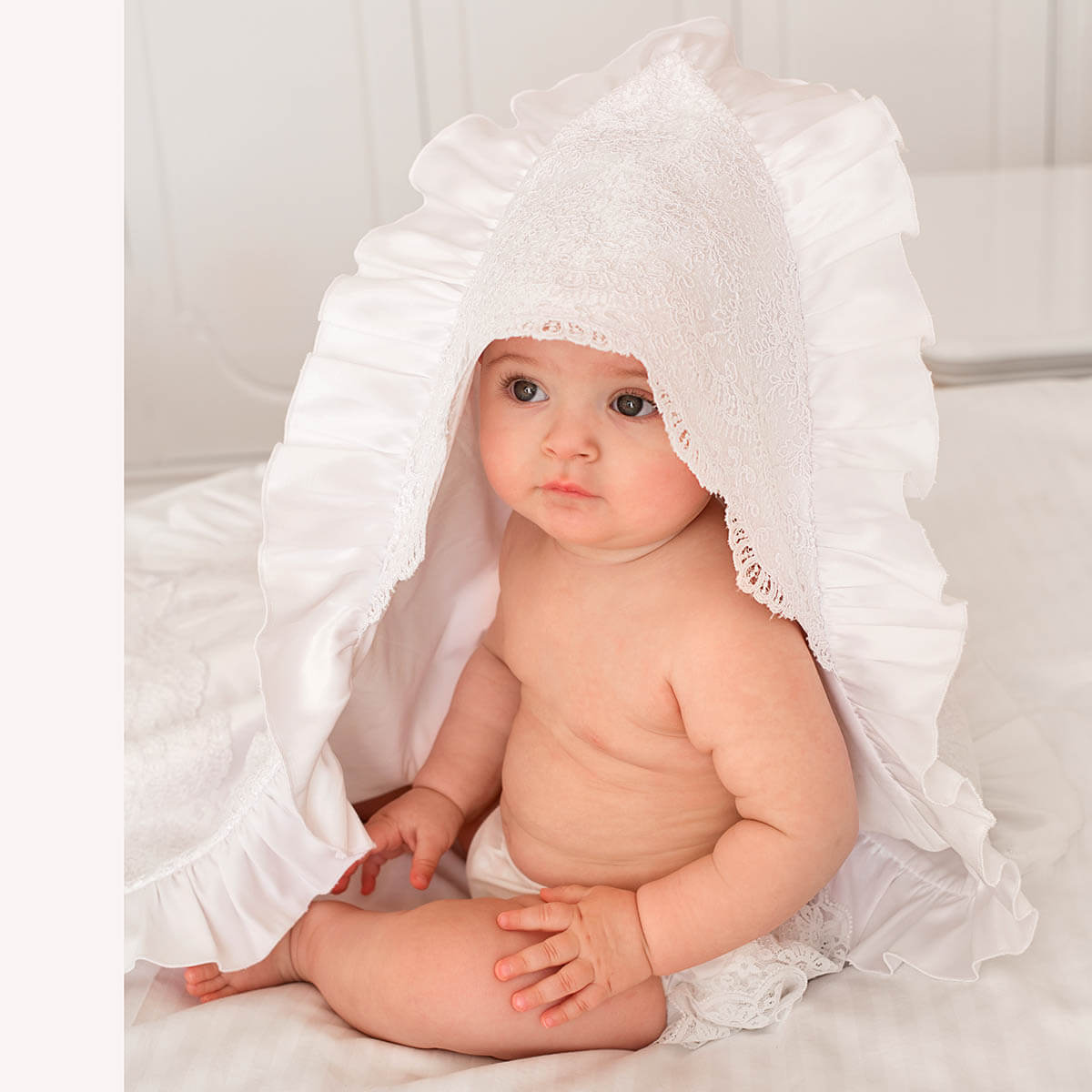 Baby wrapped in a white chrristening blanket sitting on a white surface.