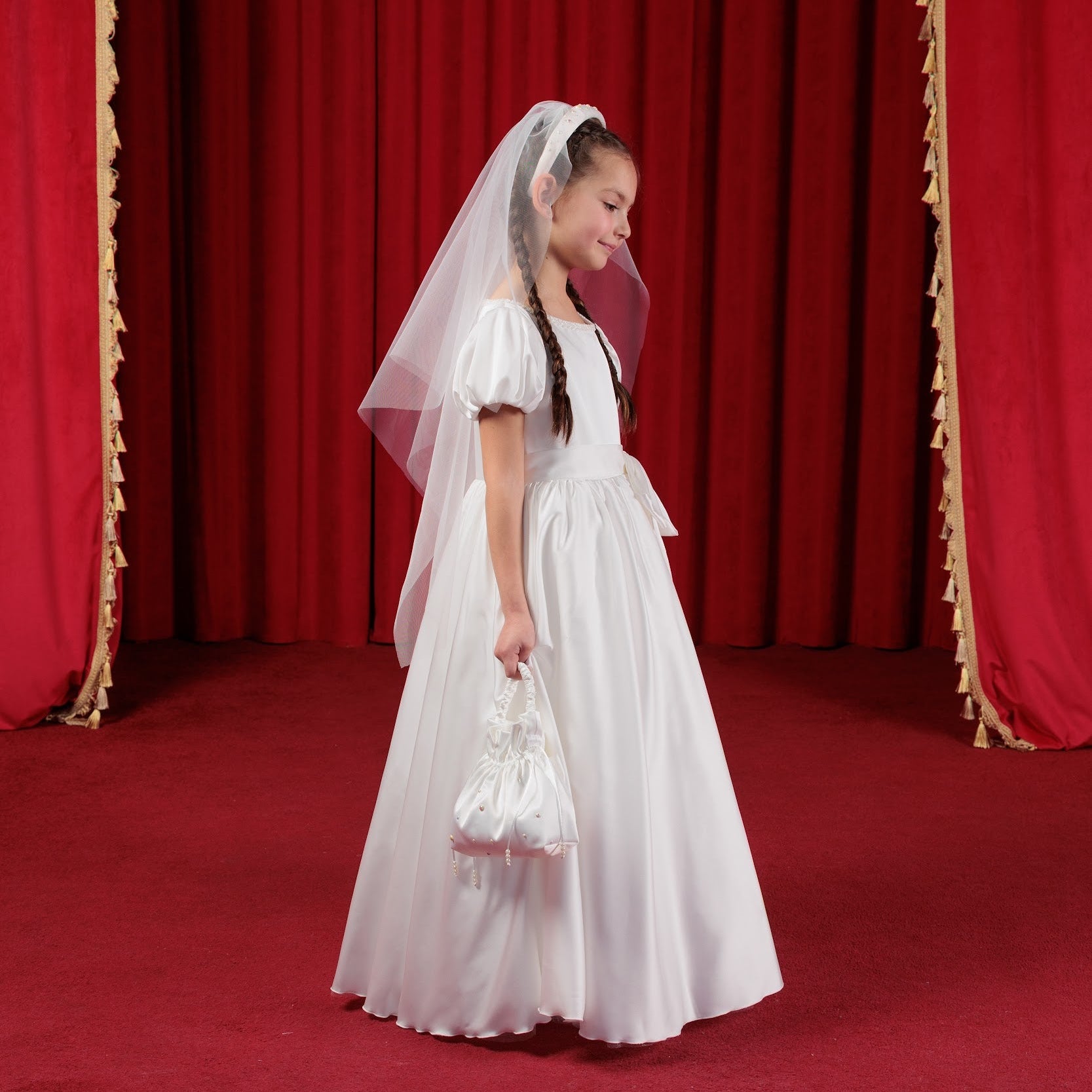 Young girl in a white dress with a veil standing in front of red curtains.