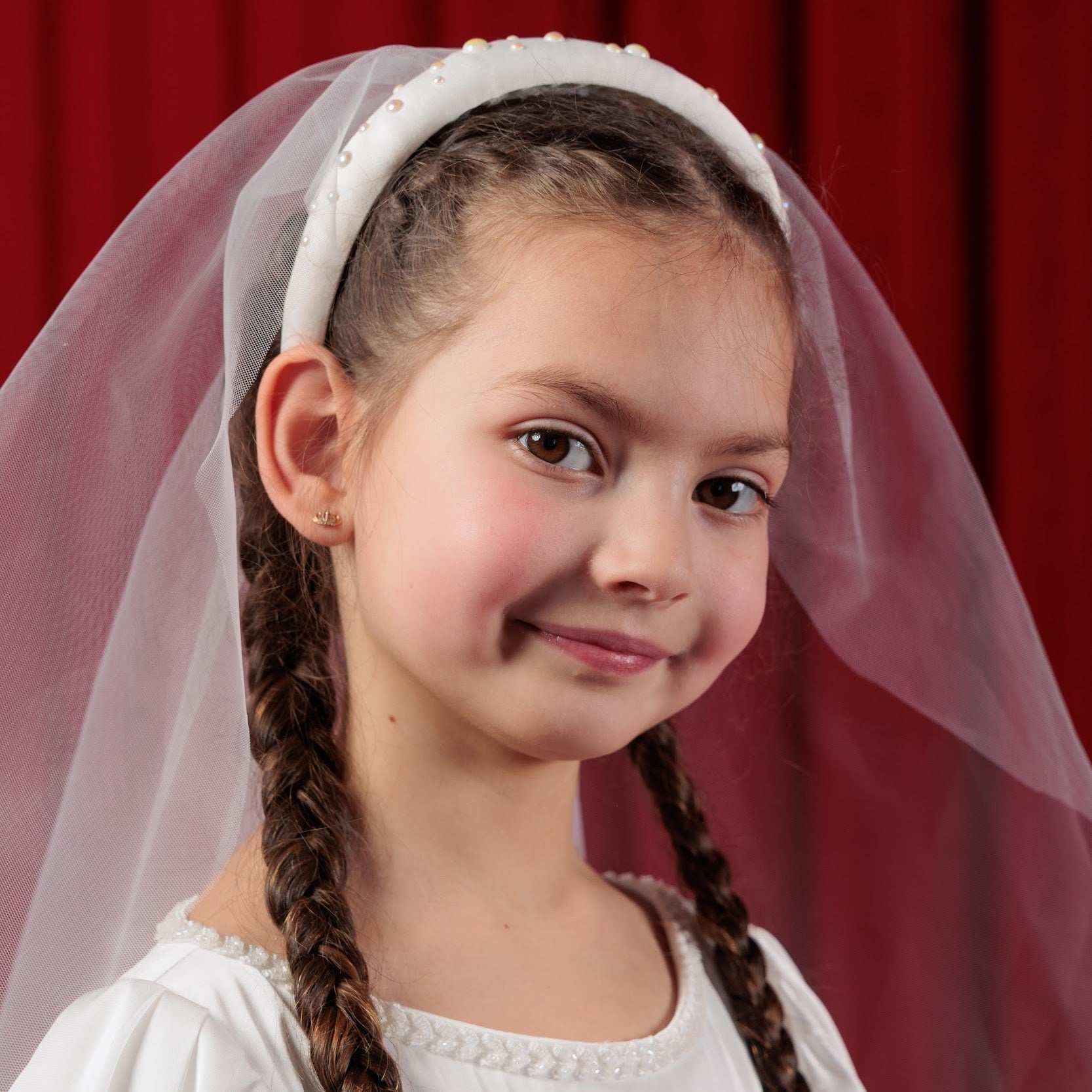 Young girl with braided hair wearing a white veil against a red curtain background