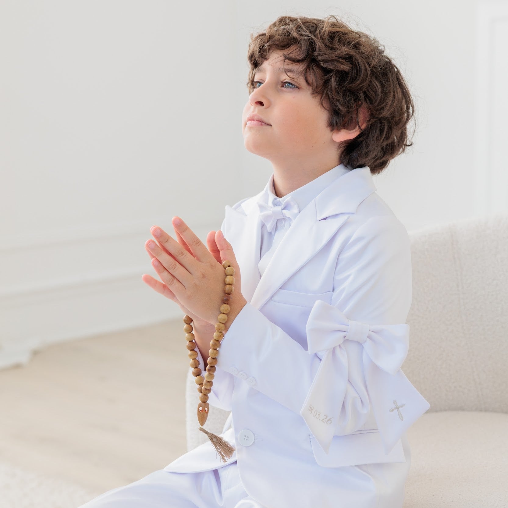 Child in white outfit with prayer beads sitting on a white couch.