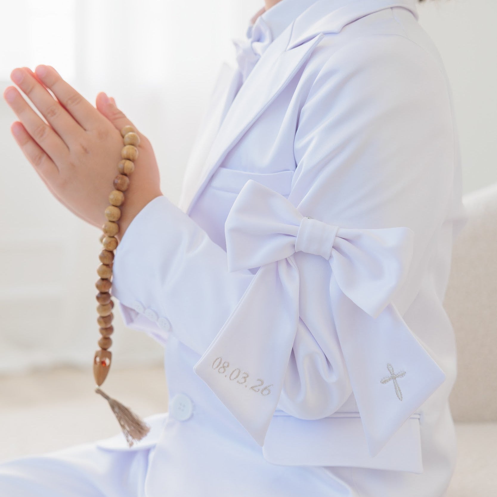 Person in light blue outfit with a bow tie, holding prayer beads on a white background