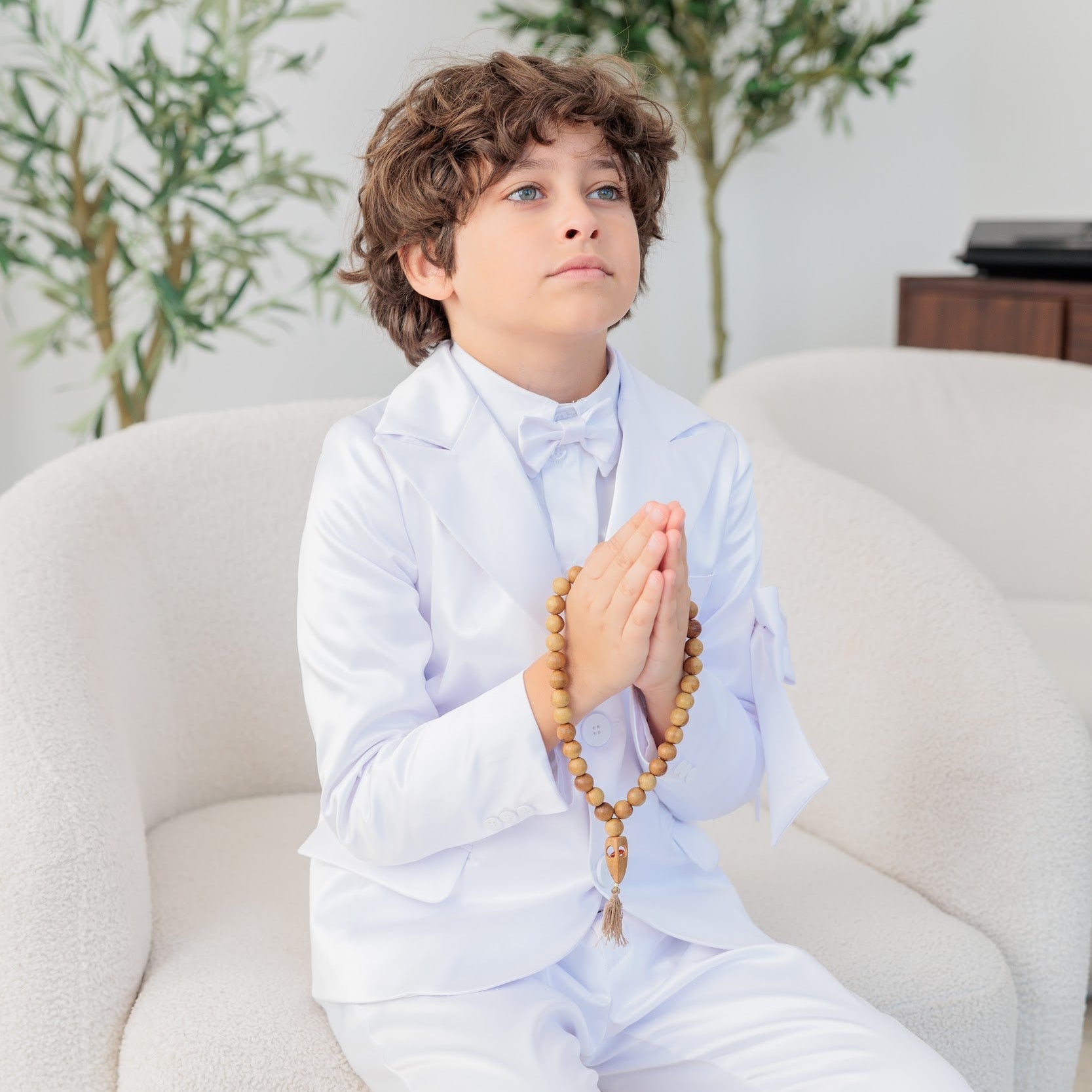Child in formal attire sitting on a white couch with prayer beads, surrounded by green plants.