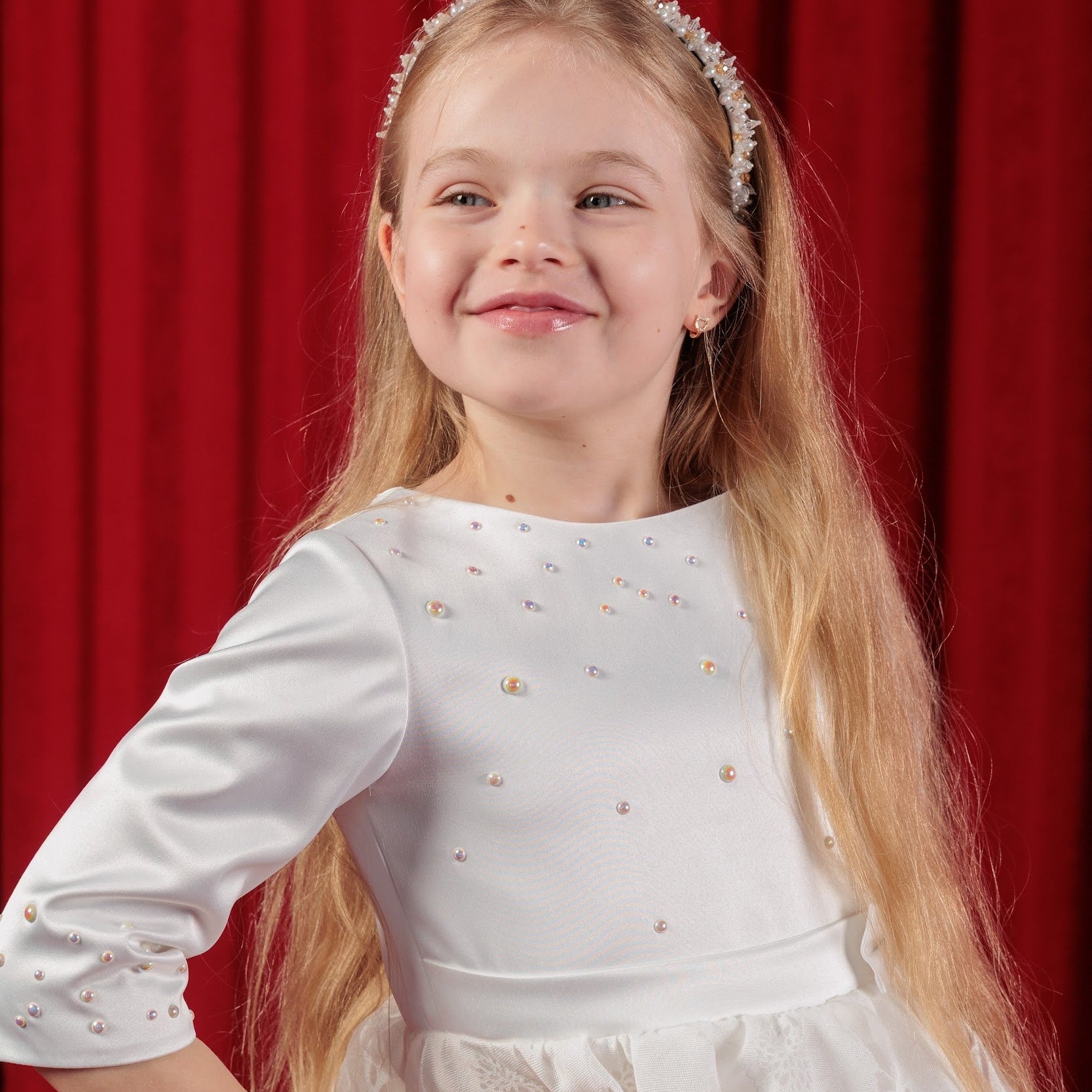 Young girl in a white dress with a headband against a red curtain background