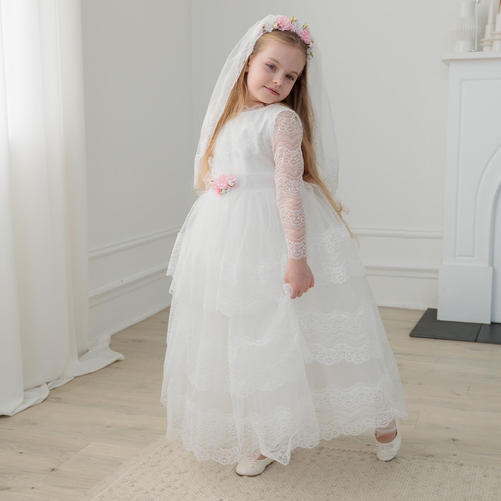 Young girl in an ivory lace communion dress with long sleeves and a veil standing in a room with white walls and a fireplace.