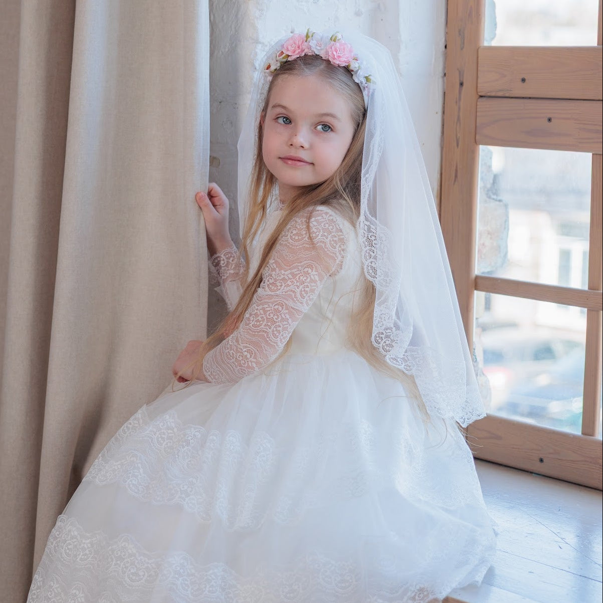 Young girl in a white lace dress standing in a room with wooden floor and large window.