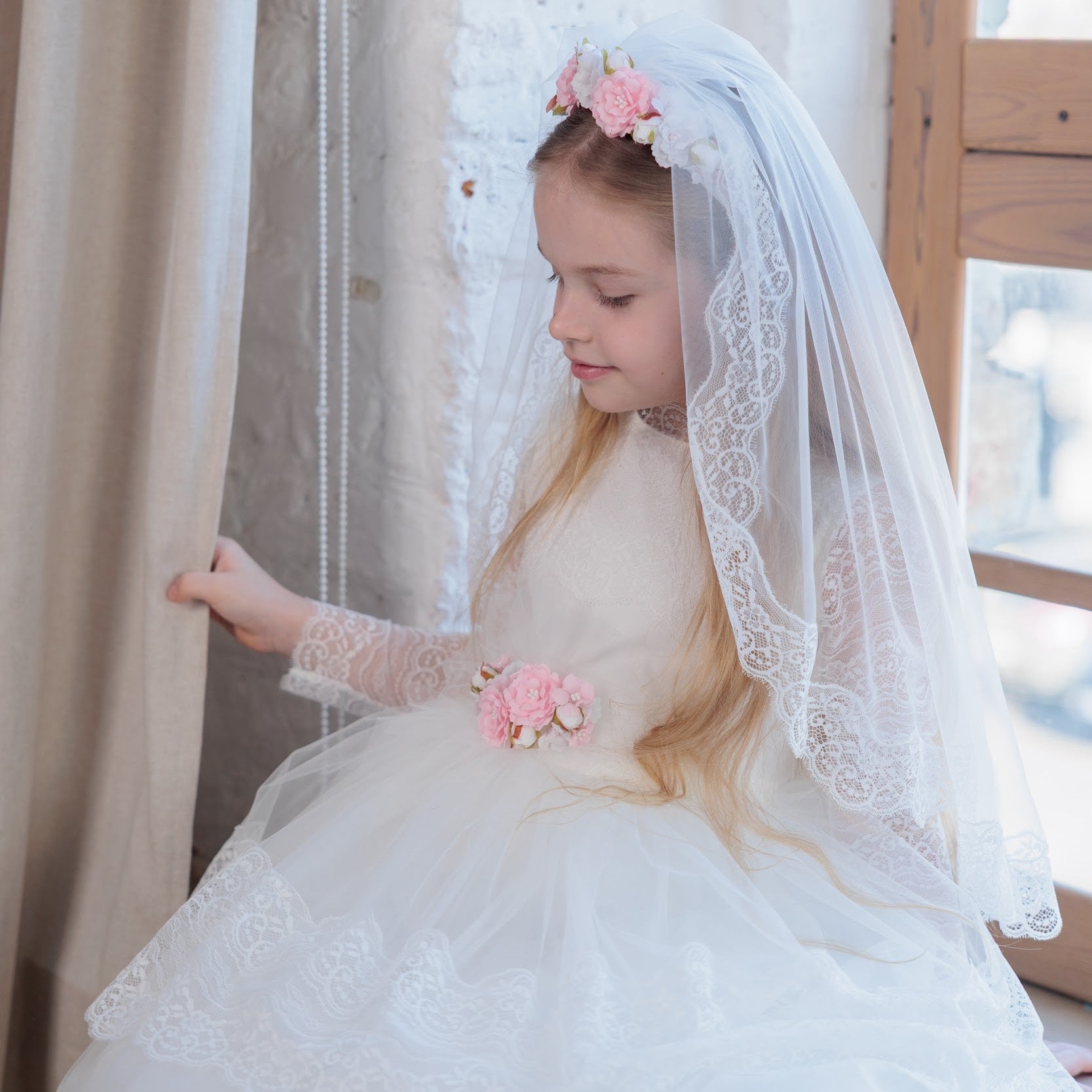 Young girl in an ivory lace communion dress with a veil and pink flowers, sitting near curtains.