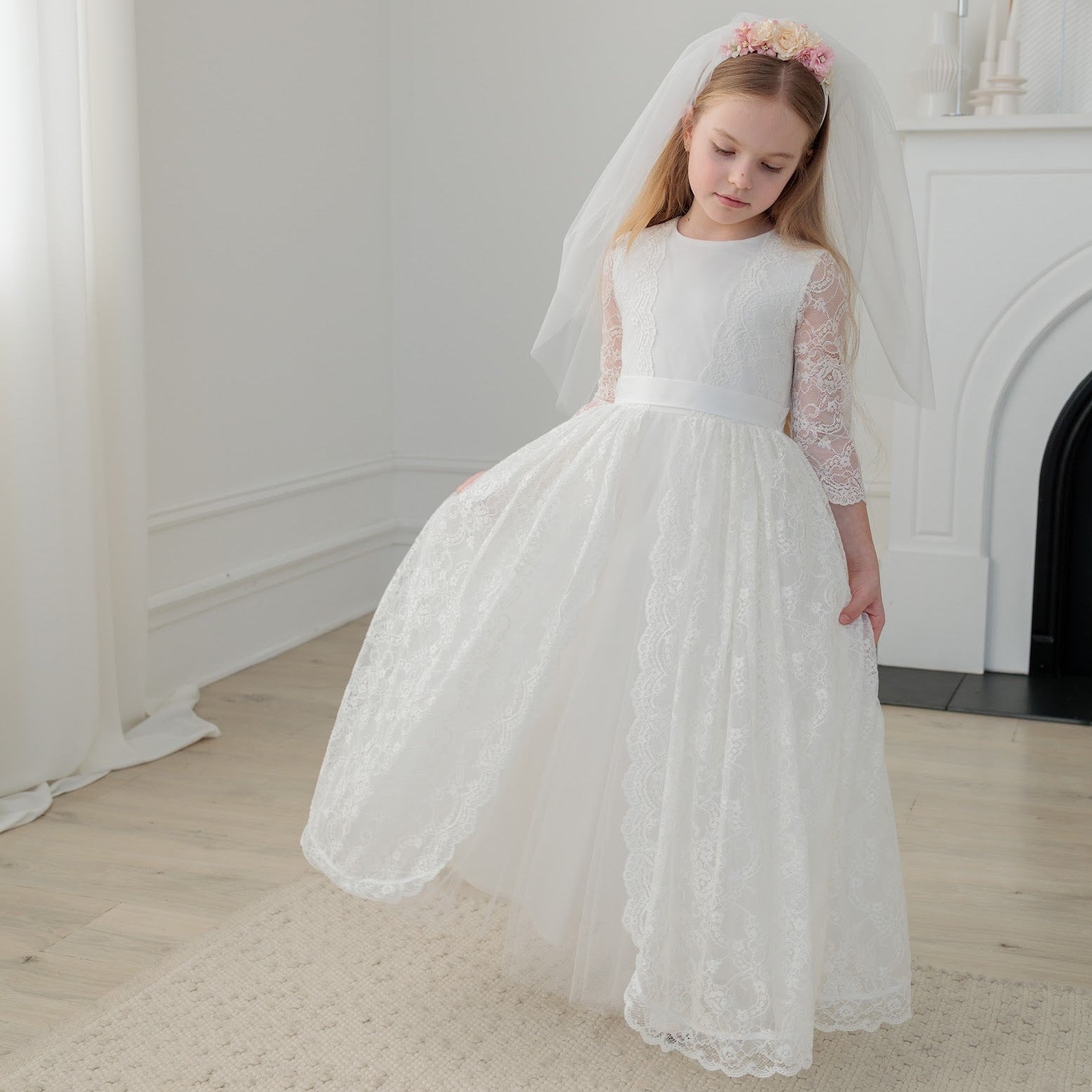 Young girl in an ivory lace communion dress with a veil standing in a room with a fireplace.