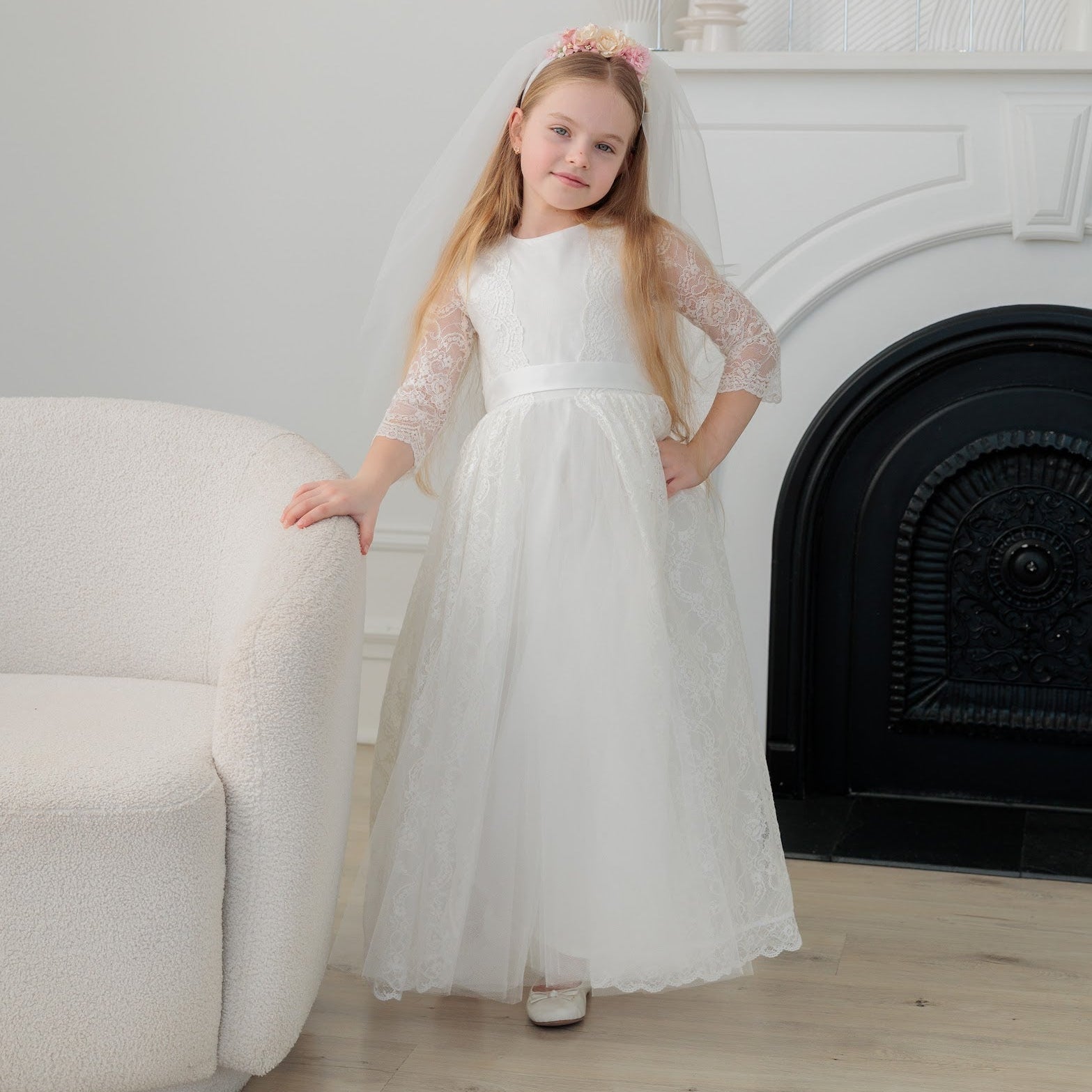 Young girl in an ivory lace communion dress standing in a room with a fireplace.