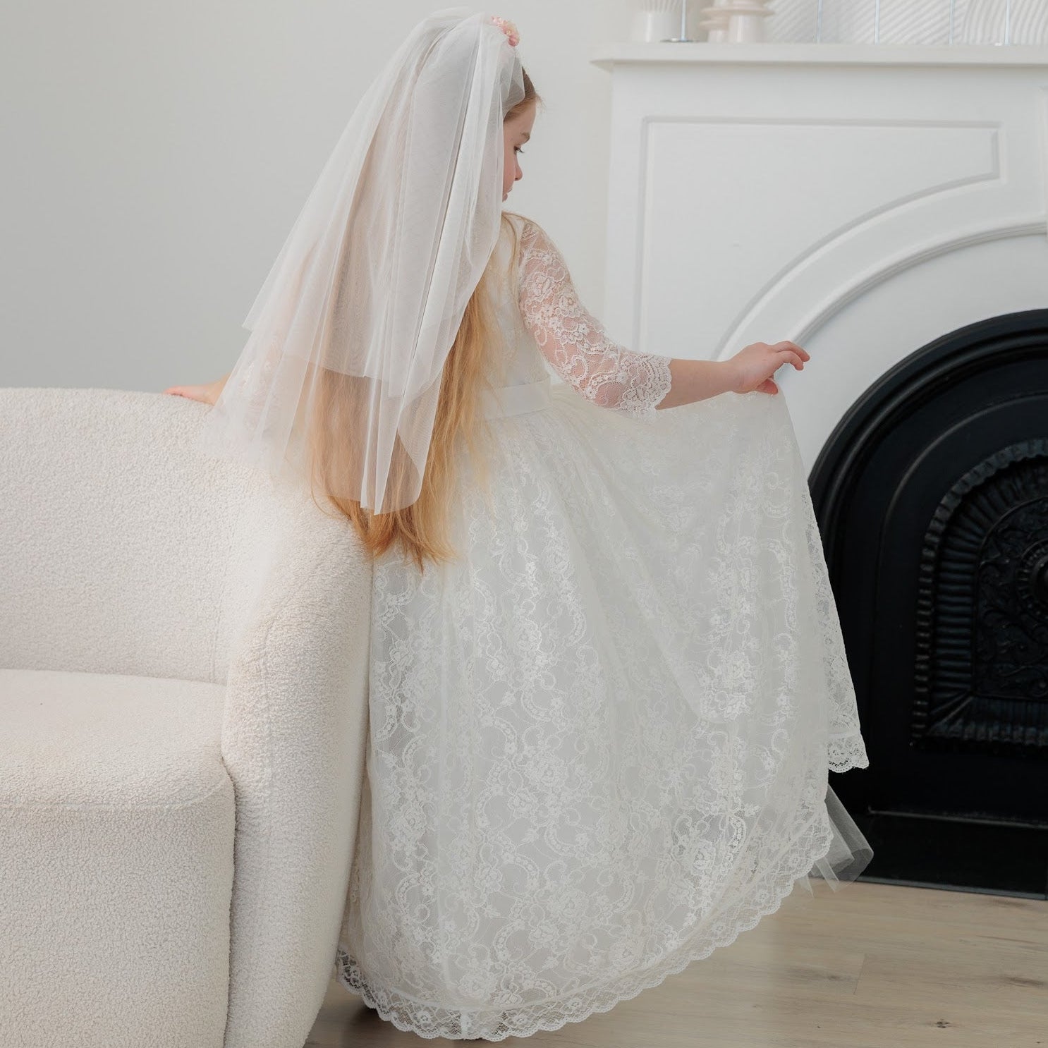 Young girl in an ivory lace communion dress with a long veil standing in a room with a fireplace.