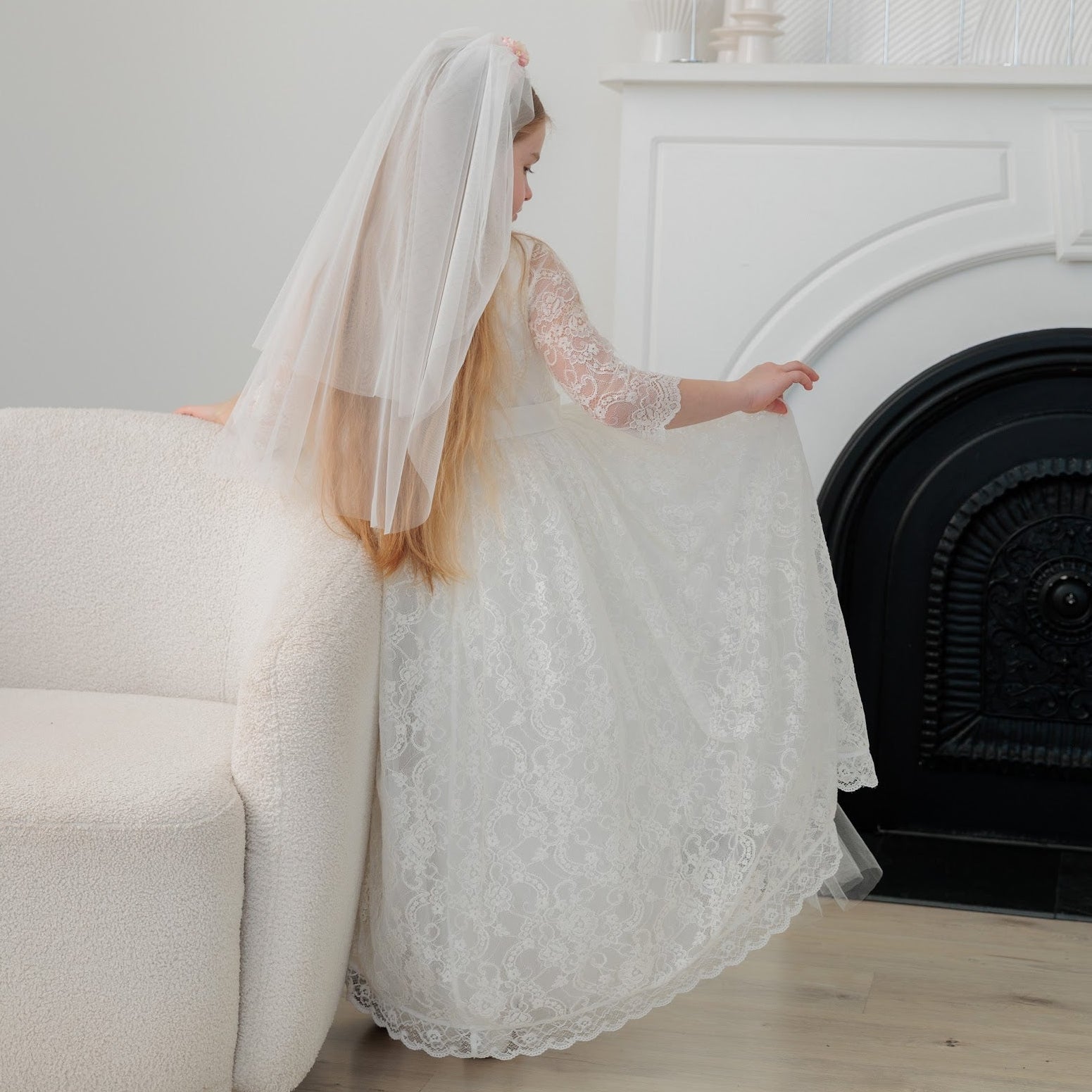 Young girl in a white lace dress with a long veil standing in a room with a fireplace.