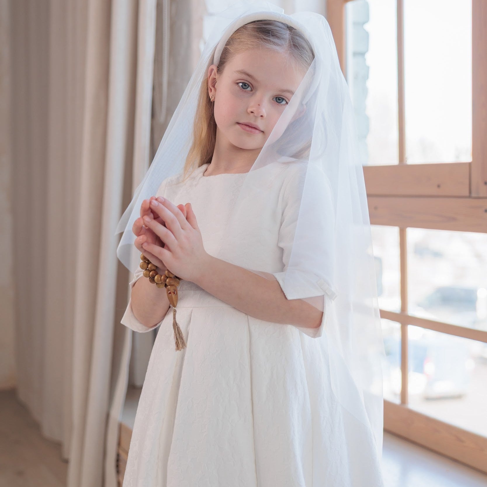 Young girl in a white communion dress with a veil standing in a softly lit room.