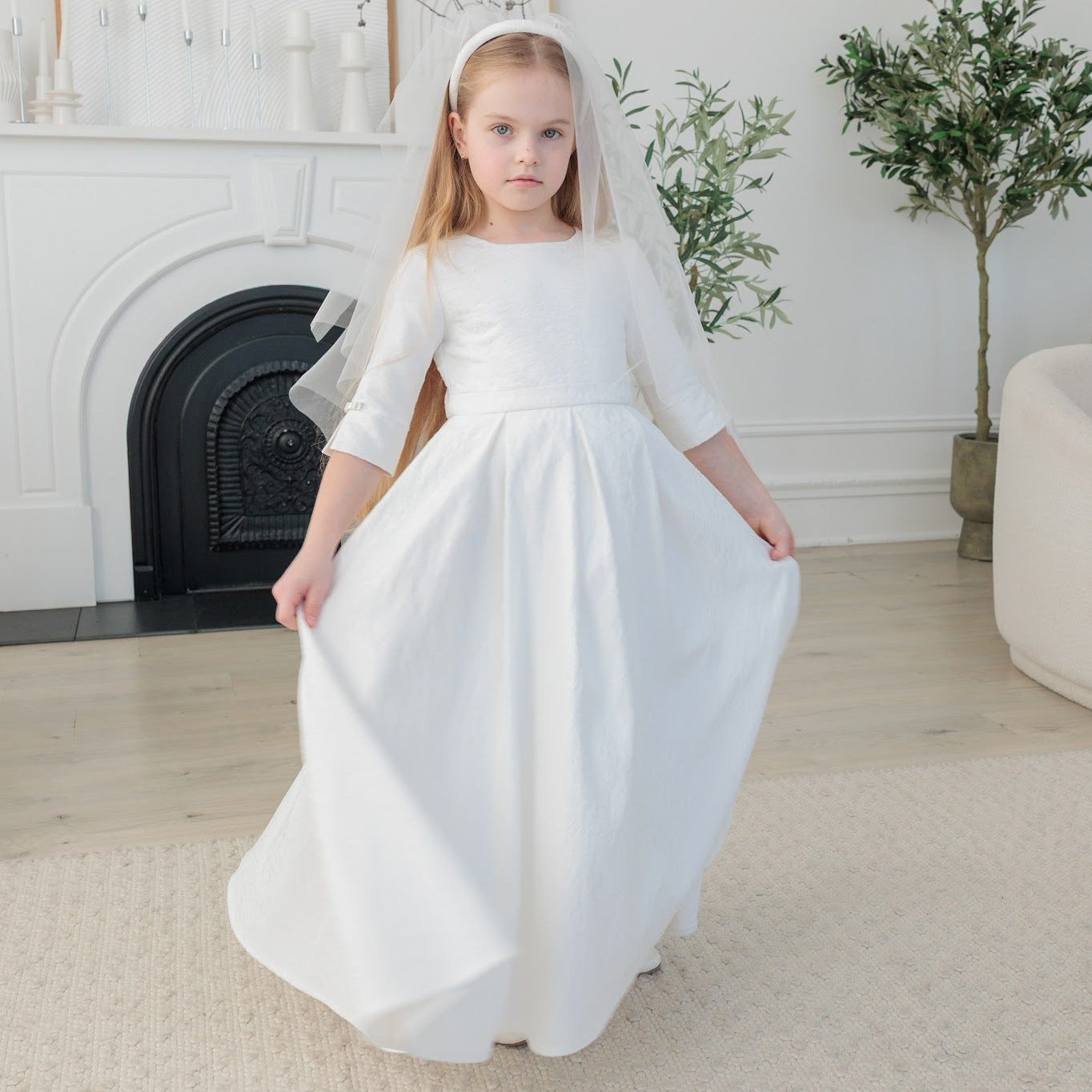 Young girl in a white communion dress standing in a room with a fireplace and plants.