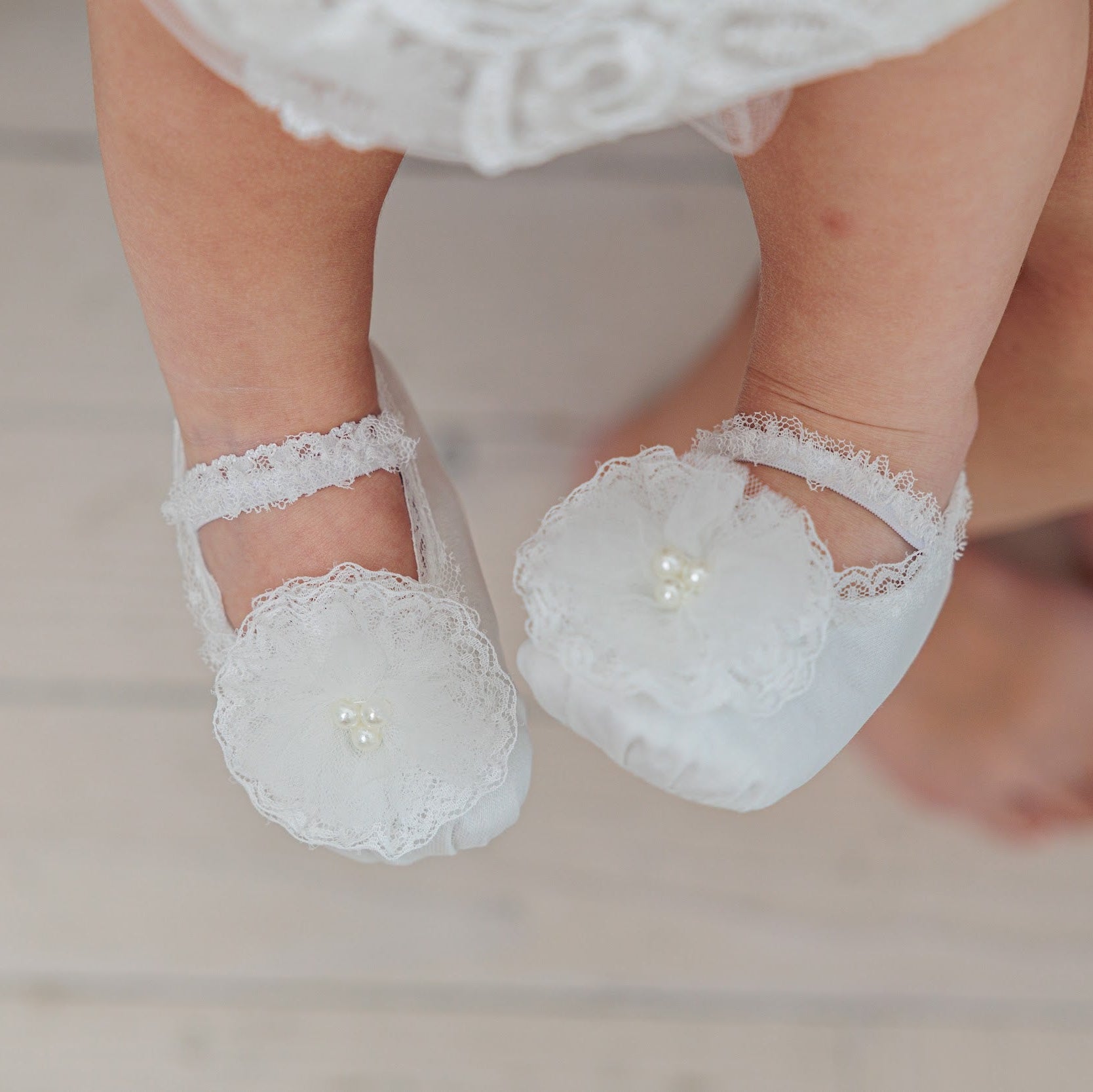 White lace baby booties with floral appliques and pearl centers, shown on a baby's feet against a light wood floor background.