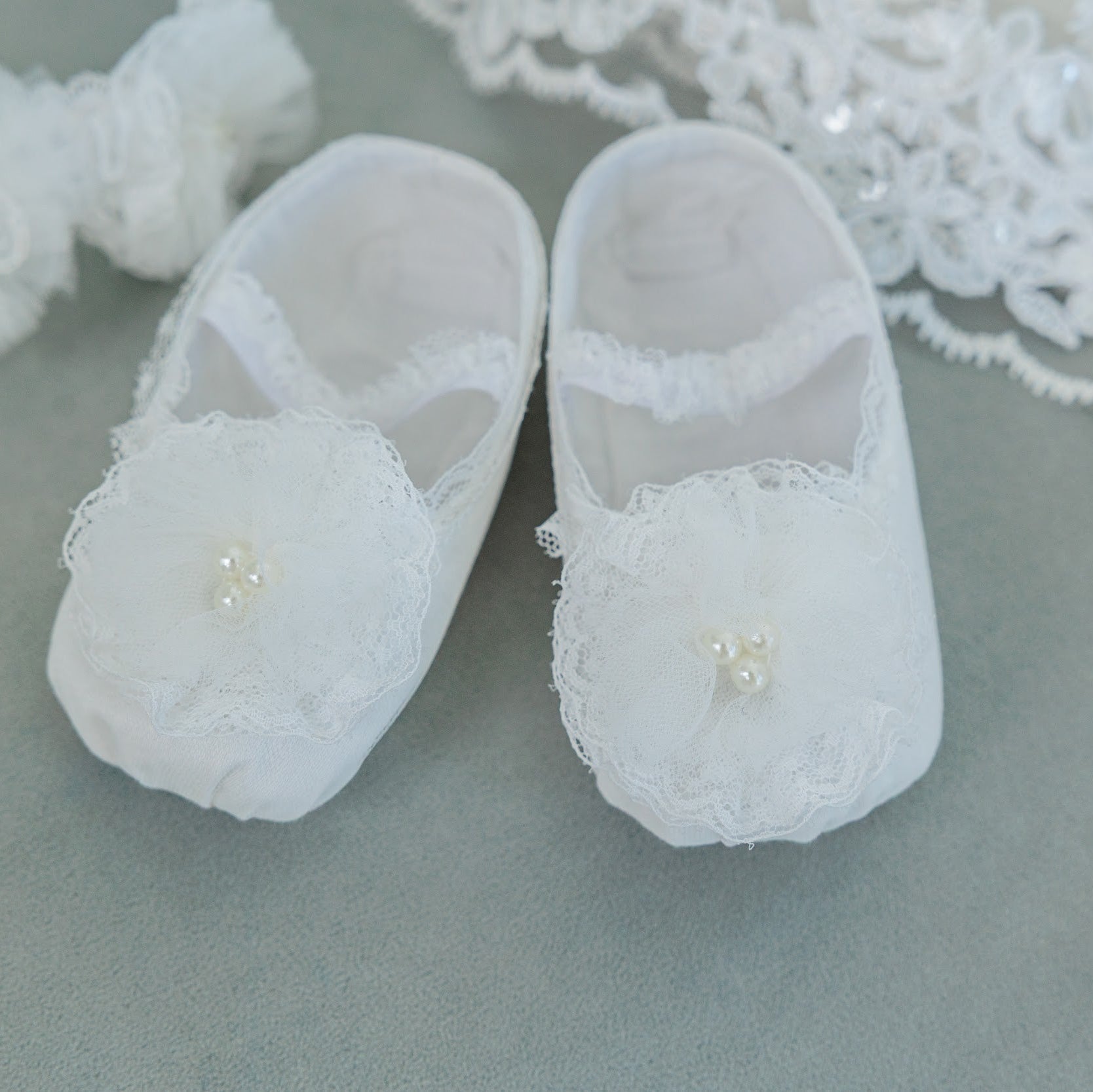 A pair of white baby booties with lace trim and pearl accents, displayed on a gray surface with decorative lace and fabric props.