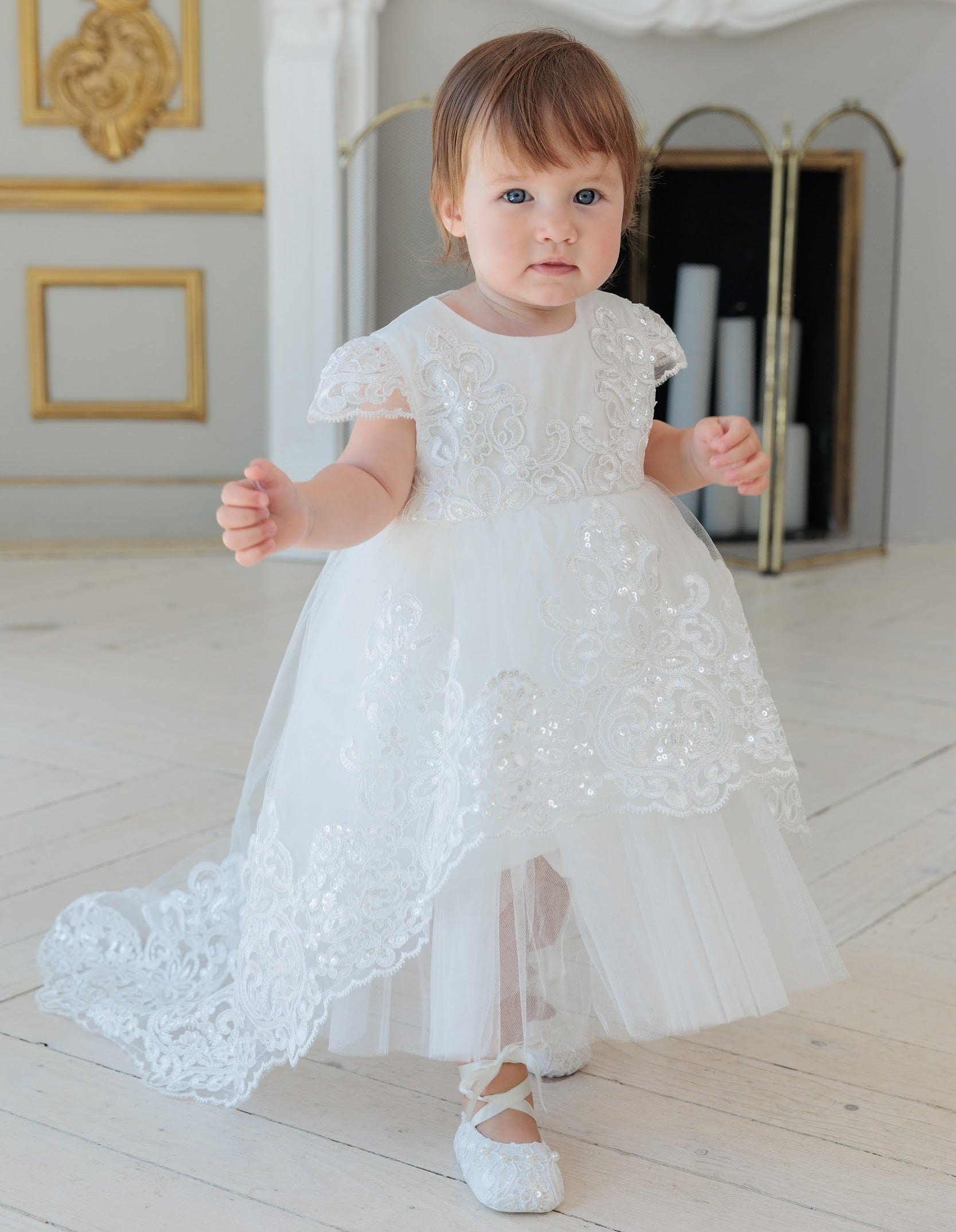 A toddler girl wearing a white lace and tulle baptism dress with short flutter sleeves and white ballet flats, standing in a bright, elegant room with a fireplace.