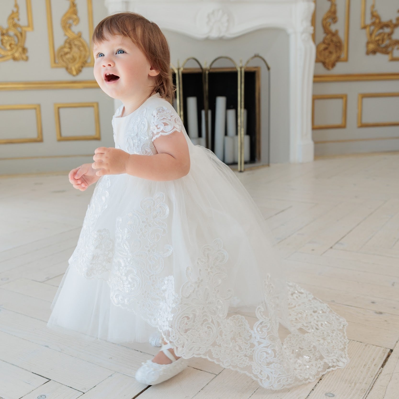 A toddler girl wearing a white lace and tulle baptism dress is captured mid-step in a bright, elegant room with gold-trimmed walls and a white fireplace.