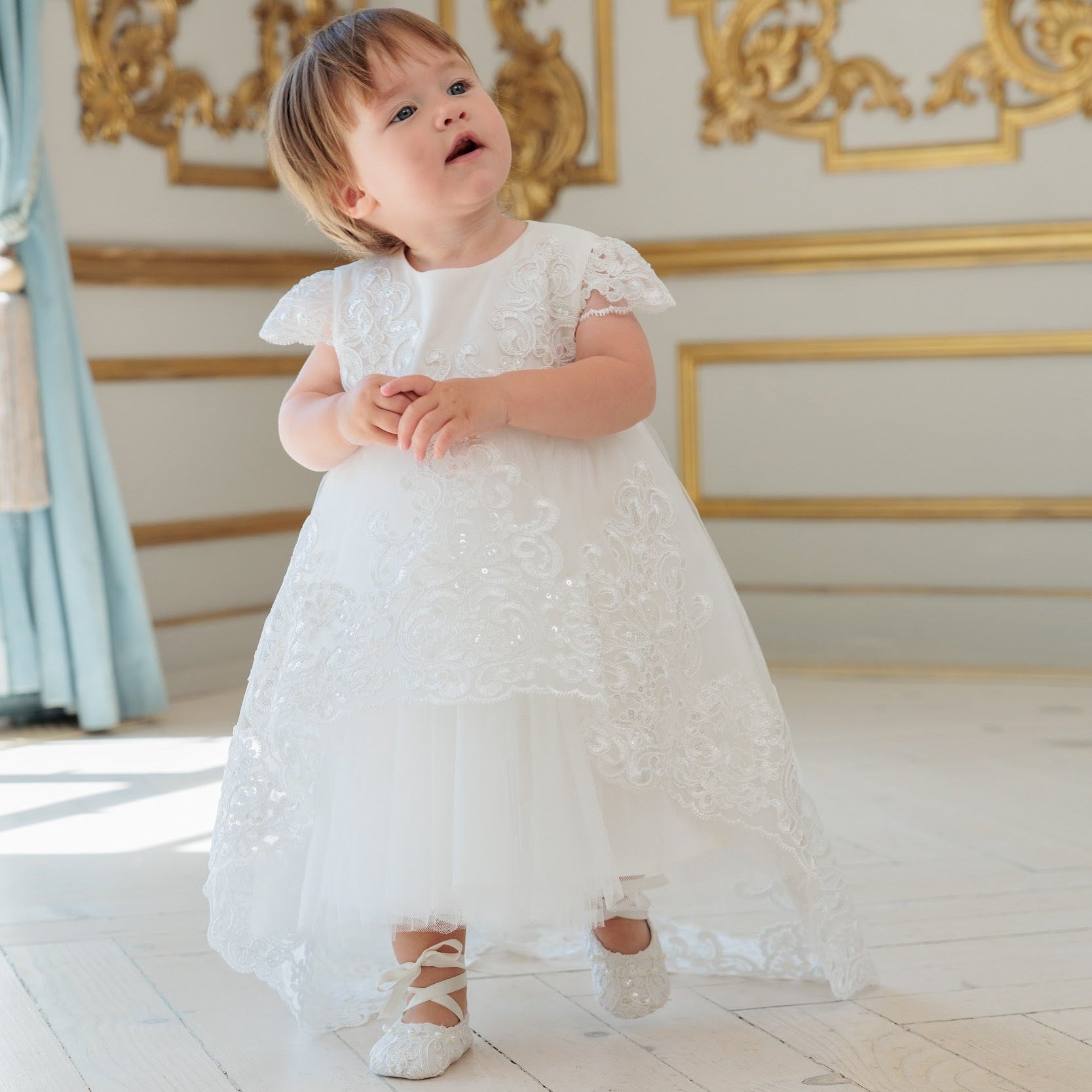 A toddler girl wearing a white lace and tulle baptism dress with lace-up shoes, standing in a grand room with gold wall decorations and light wood floors.