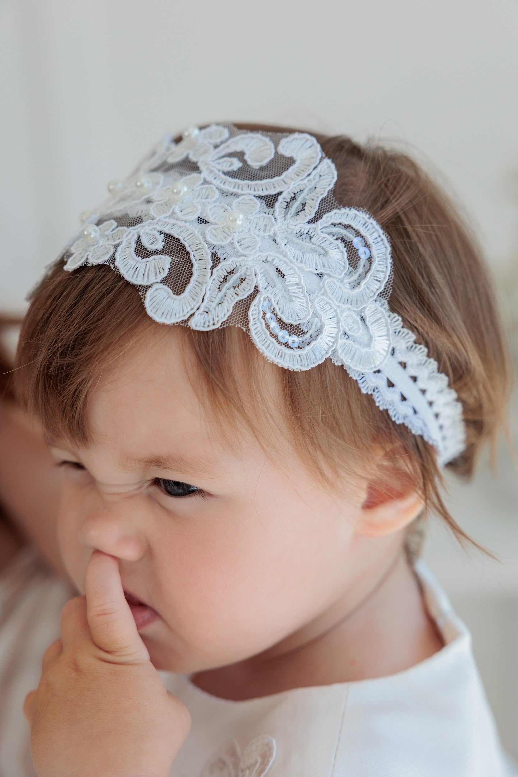 A close-up of a young child wearing a white lace headband with floral embroidery and small pearl-like accents, photographed against a soft light background.