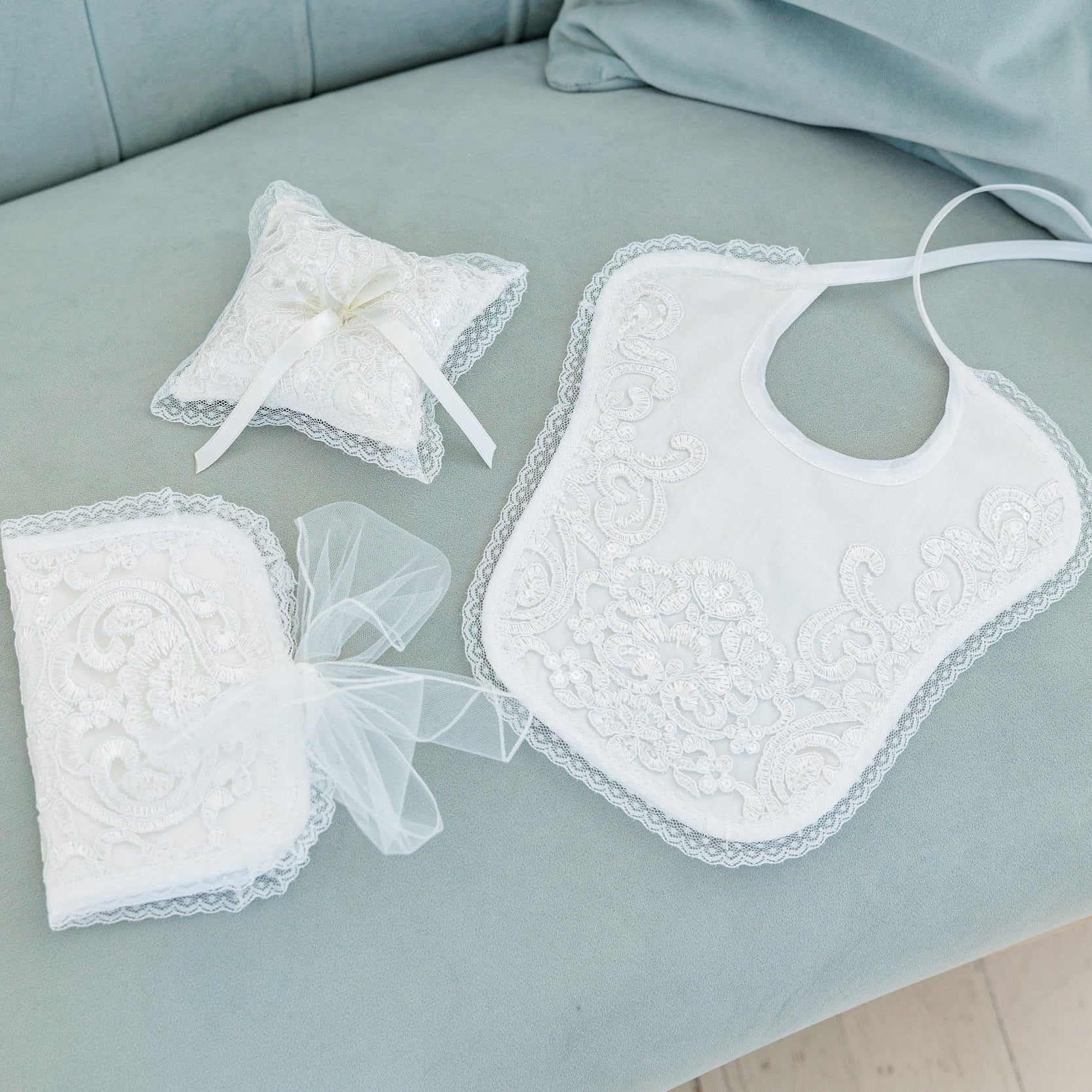 White lace baby gift set including a bib, ring pillow, and small gift bag with sheer ribbon, arranged on a light blue-green sofa.