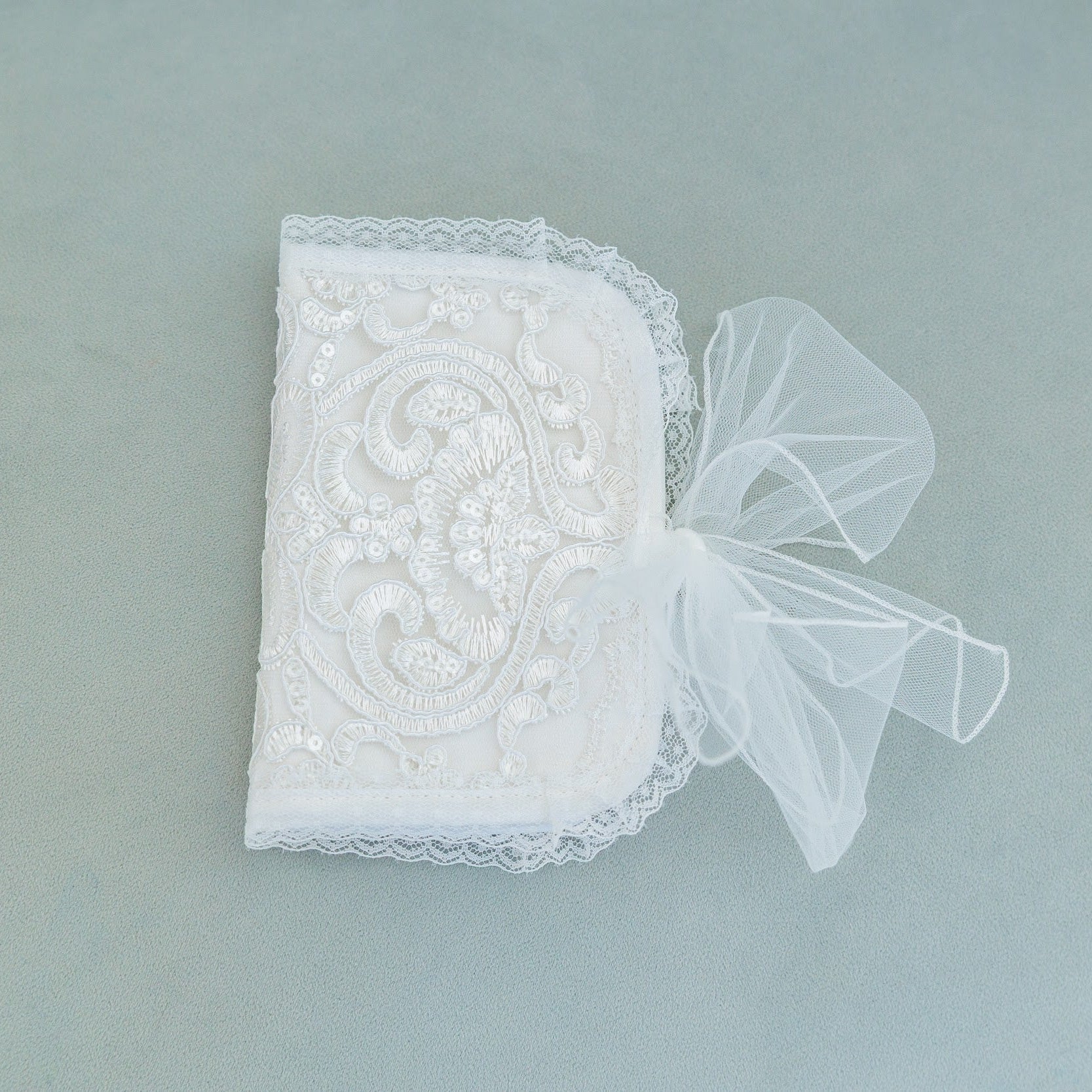 A white embroidered lace NICA Baby First Curl Keepsake Box with a sheer white tulle bow on top, displayed on a muted gray-green background.