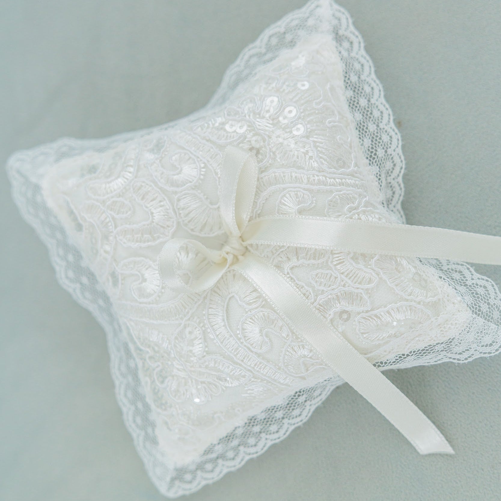 An ivory lace ring pillow with a satin ribbon bow, displayed on a soft, pale sage green background.