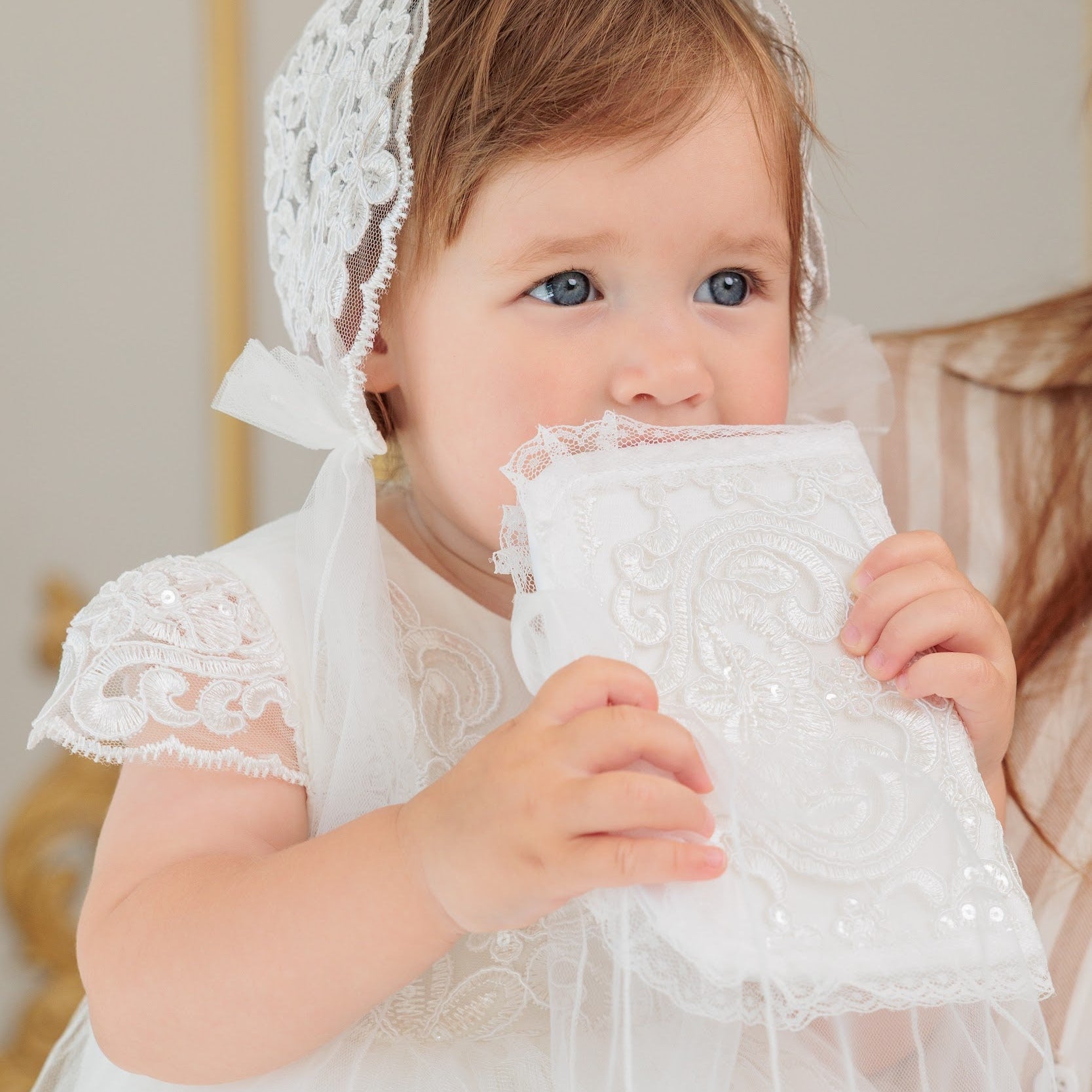 A baby girl wearing a white lace first communion dress and matching lace bonnet holds a matching white lace communion book in front of her face.