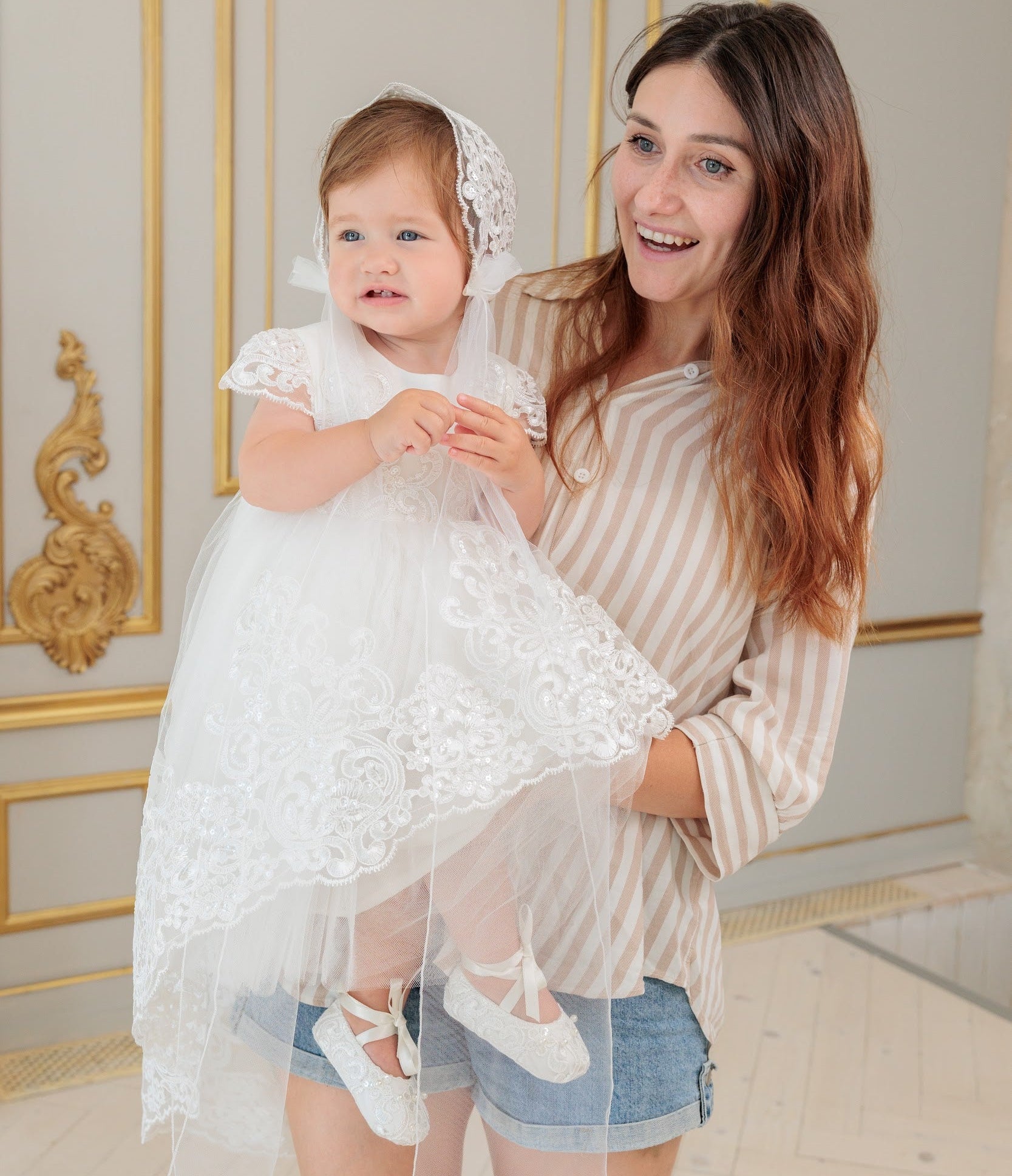 A woman holding a baby girl wearing a white lace first communion dress with a matching veil and headpiece, set against an elegant interior with gold-trimmed walls.