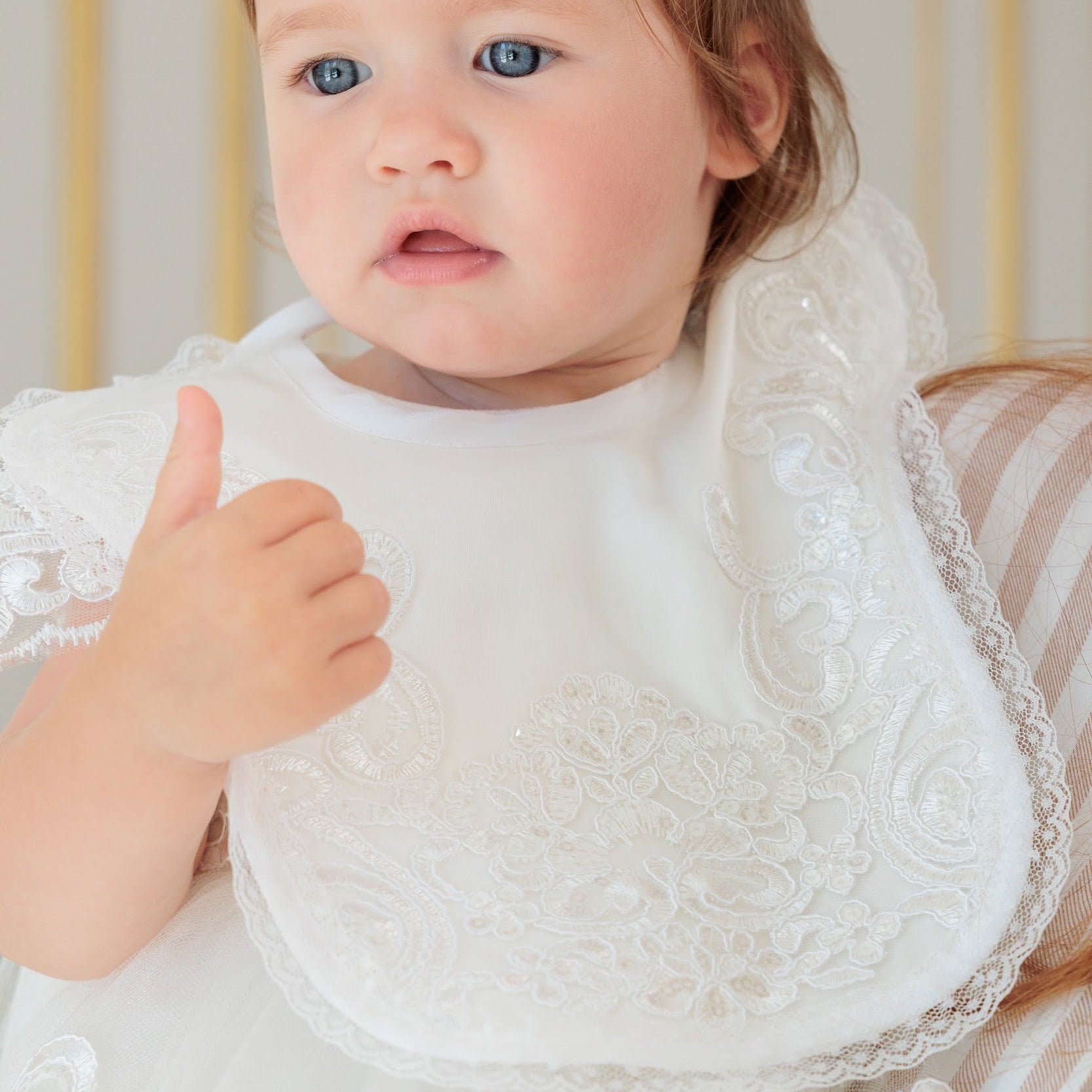 A close-up of a baby wearing a white lace bib and matching white dress, giving a thumbs-up gesture against a soft cream-colored background.