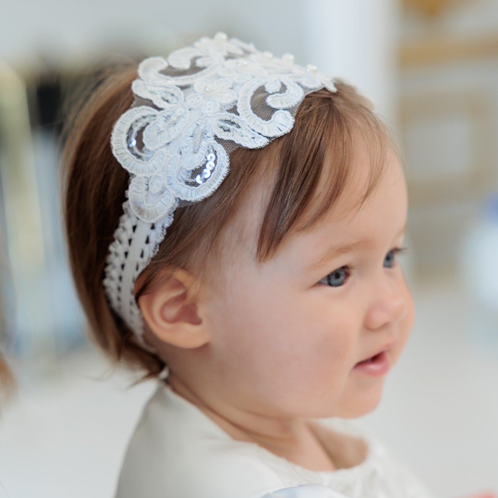 A close-up of a young child wearing a white lace headband with sequins and a white dress, set against a softly blurred indoor background.