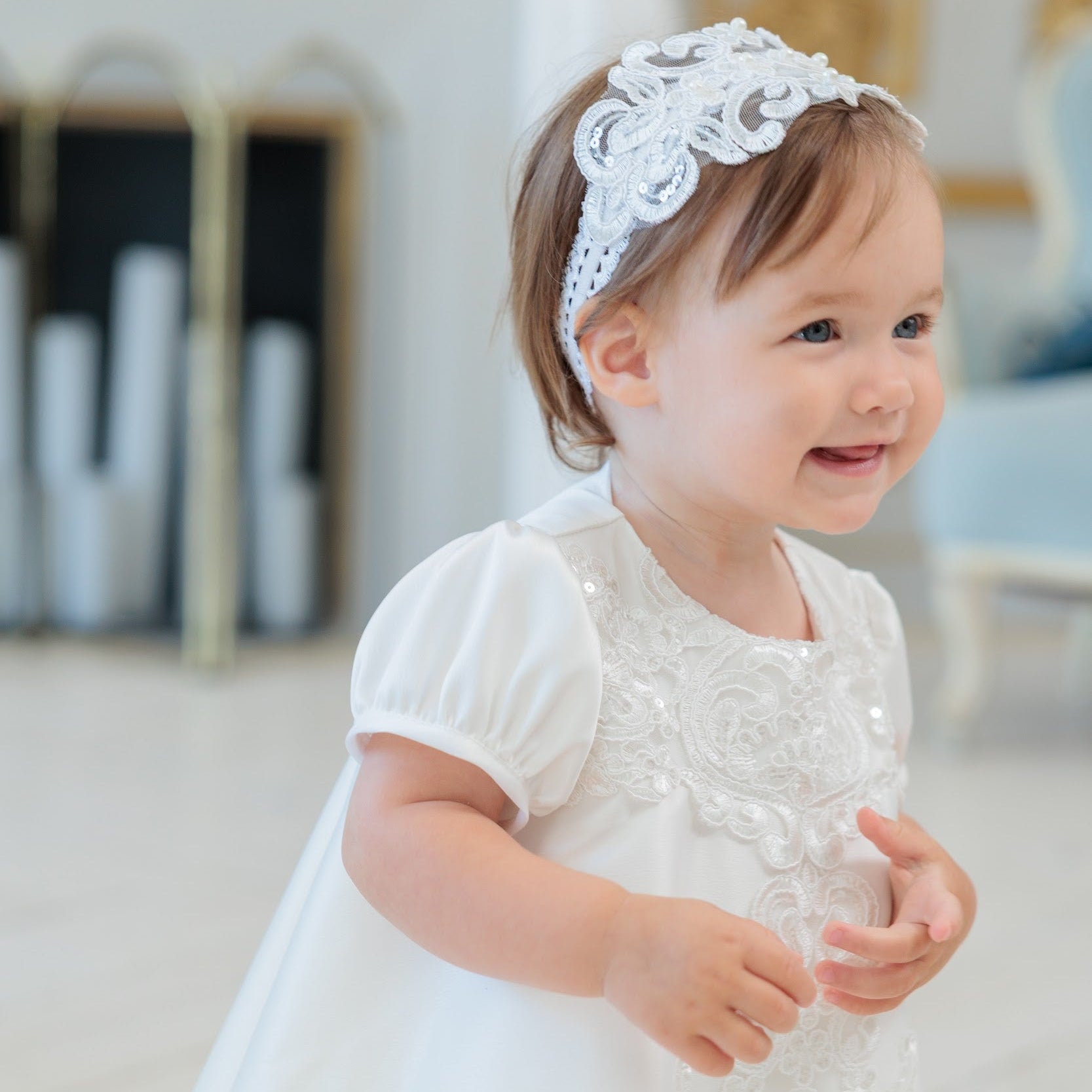 A toddler girl wearing a white lace dress with lace embroidery and a matching white lace headband, standing in a bright indoor setting.