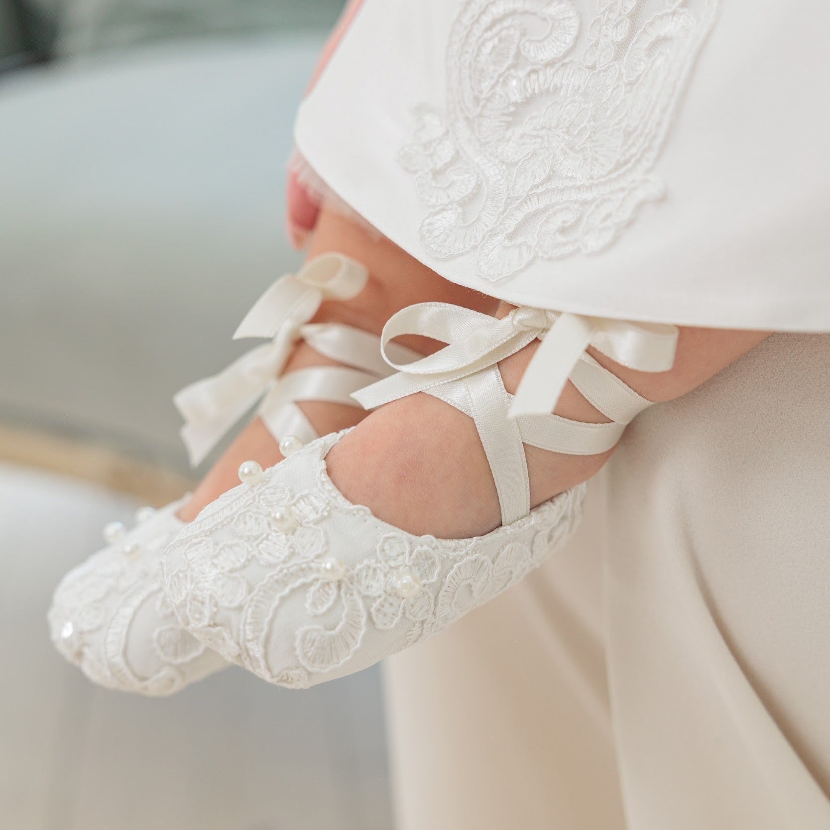 White lace baby shoes with ribbon laces and a matching white embroidered dress, shown on a baby's feet in a soft indoor setting.