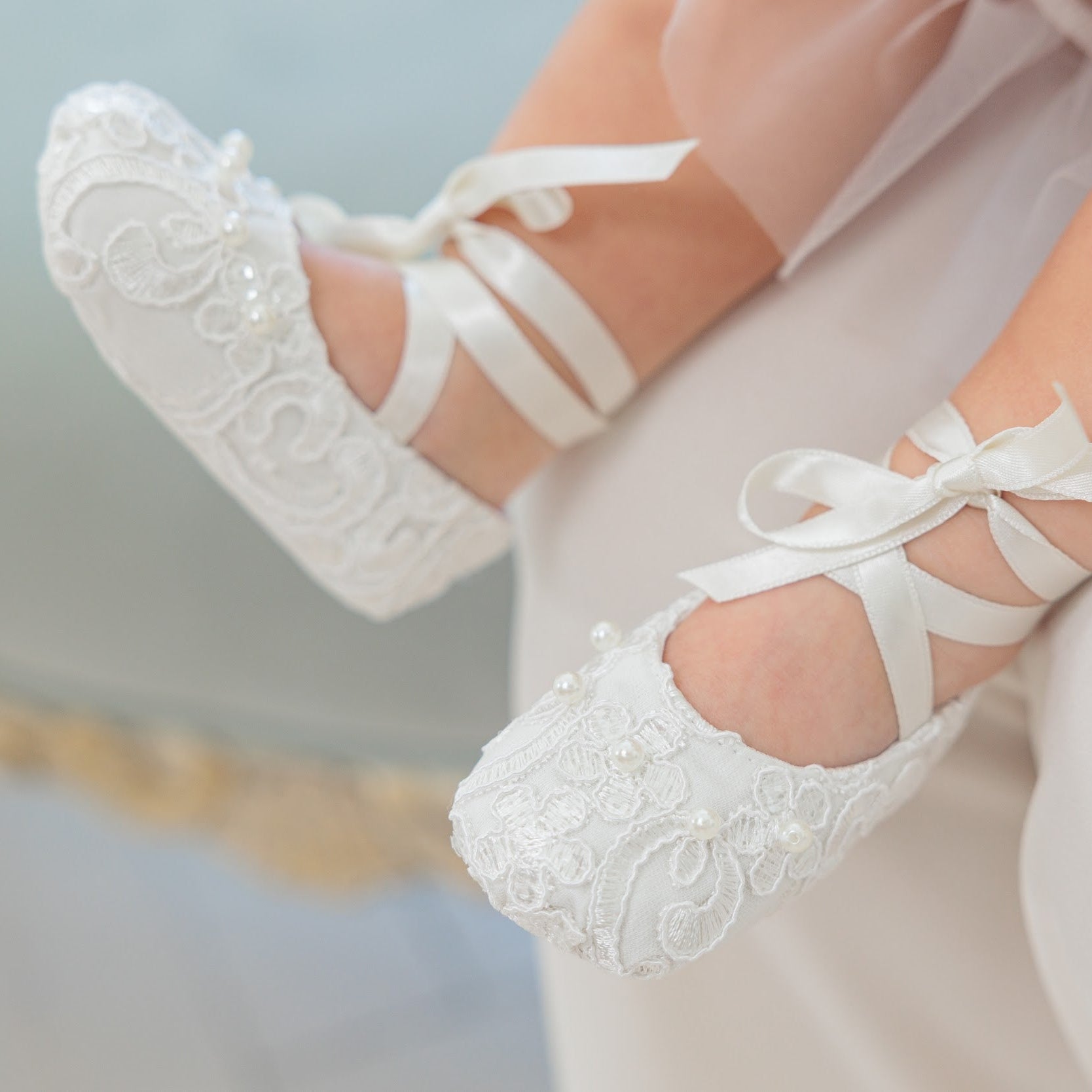 White lace baby ballet flats with ribbon bows, shown on a child's feet against a soft light background.