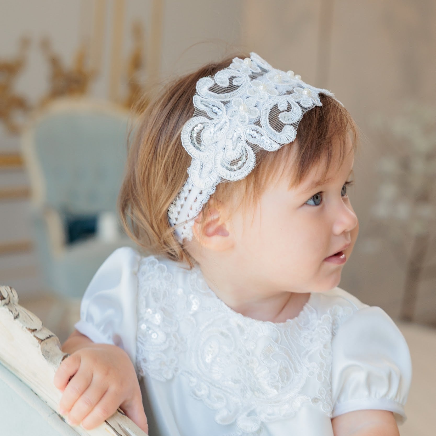 A toddler girl in a white lace dress wearing a white lace headband, looking to the side in a softly lit indoor setting.