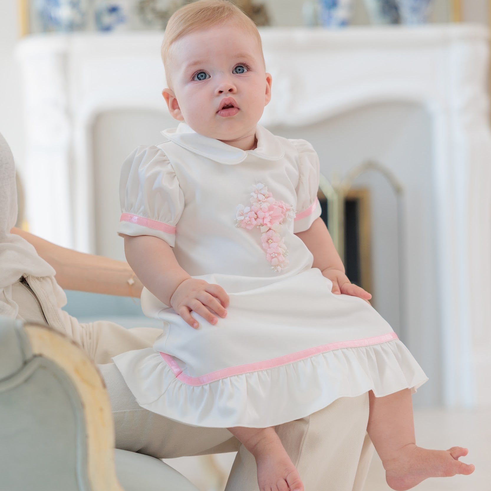 A baby girl wearing a white short-sleeve dress with pink trim and a pink floral applique, sitting on her mother's lap in a bright, elegant room.