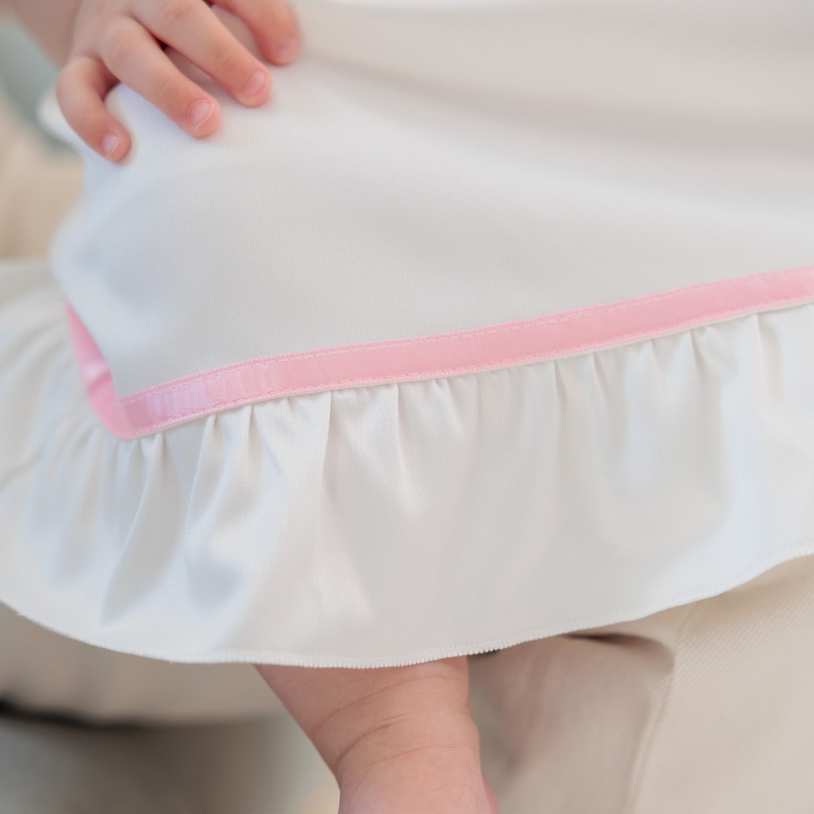 Close-up of a white baby dress or skirt with a pink ruffle trim, being worn by a baby with one foot visible and a hand resting on the fabric.