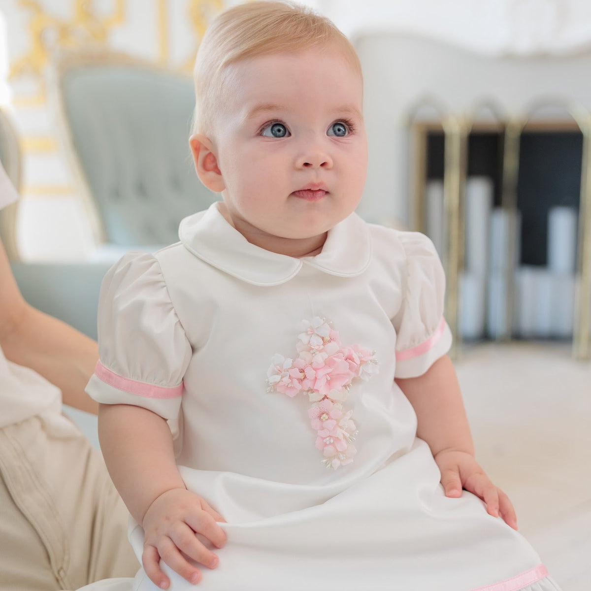 A baby girl wearing a white short-sleeve dress with pink trim and a pink floral cross on the chest, sitting on a person's lap in a bright, elegant room.