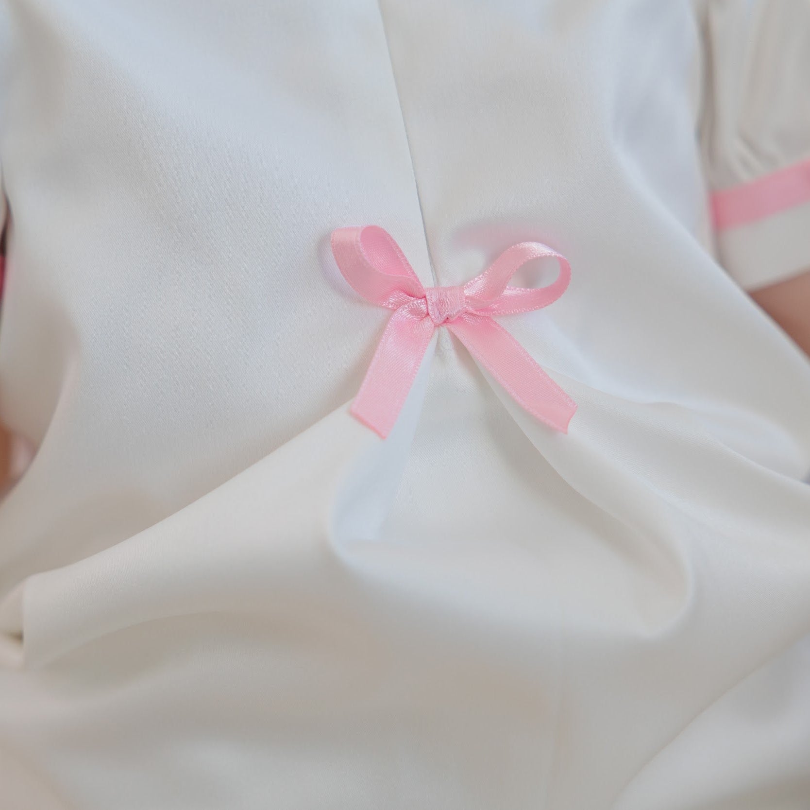 Close-up of a white satin-like garment with short puff sleeves trimmed in pink ribbon and a pink bow at the back, worn by a child.