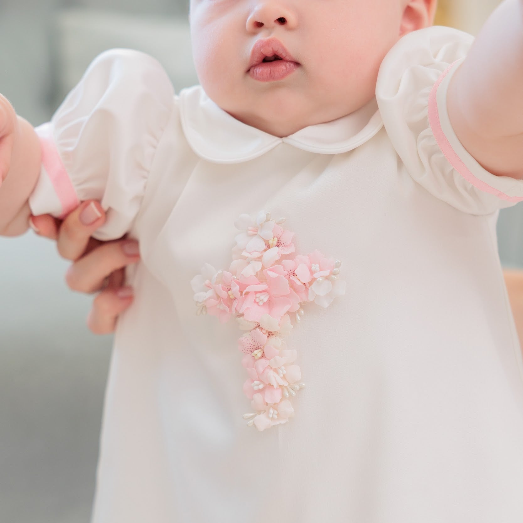 A close-up of a baby wearing a white long-sleeve dress with a Peter Pan collar and a pink floral cross applique on the chest.