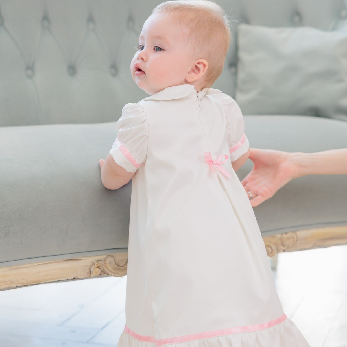A baby wearing a short-sleeved white baptism dress with pink trim and a small pink bow on the back, standing on a light wood floor next to a tufted gray sofa while an adult hand supports them.