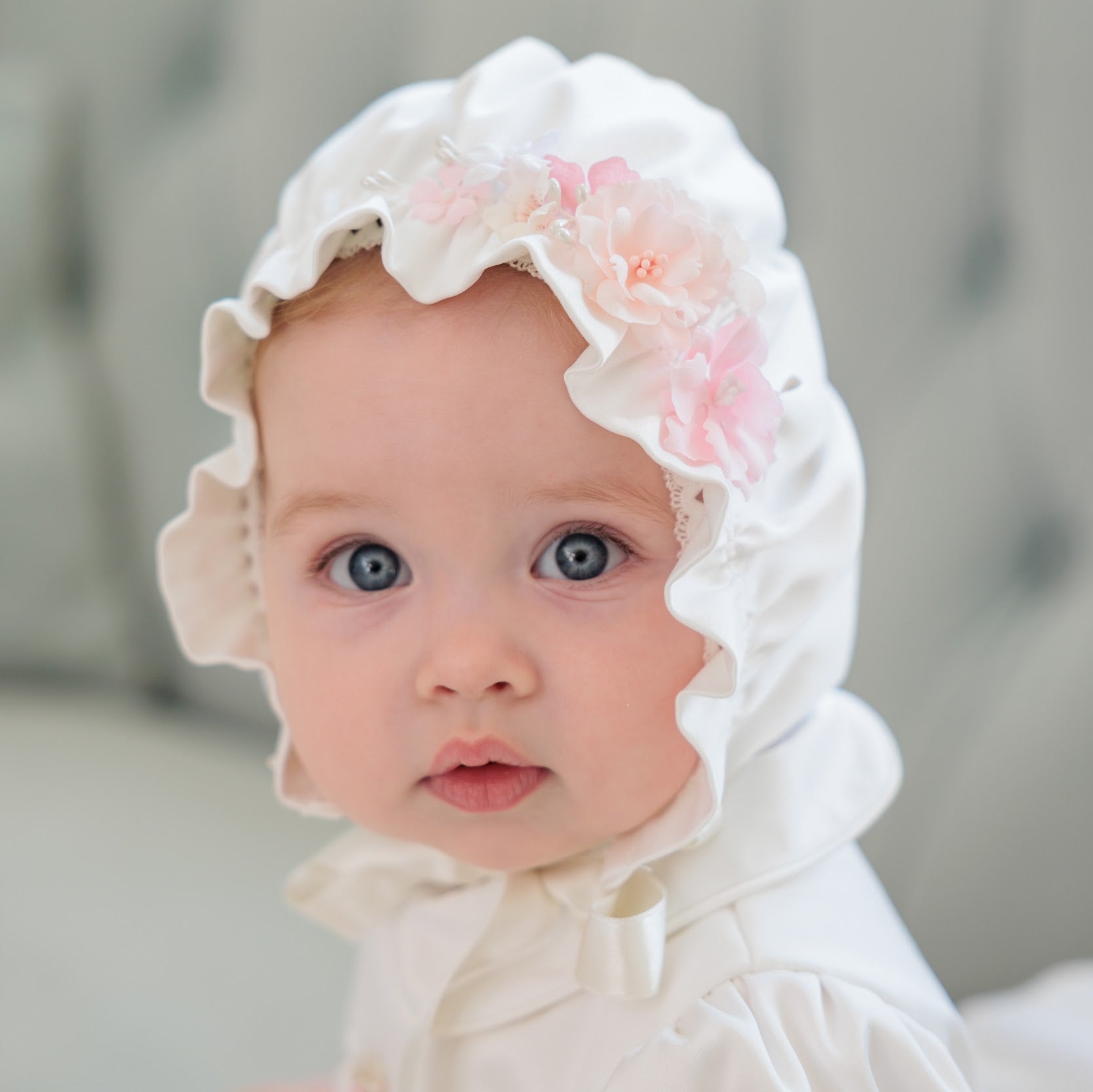 A close-up portrait of a baby wearing a white bonnet with pink fabric flowers, set against a soft, blurred gray tufted headboard.