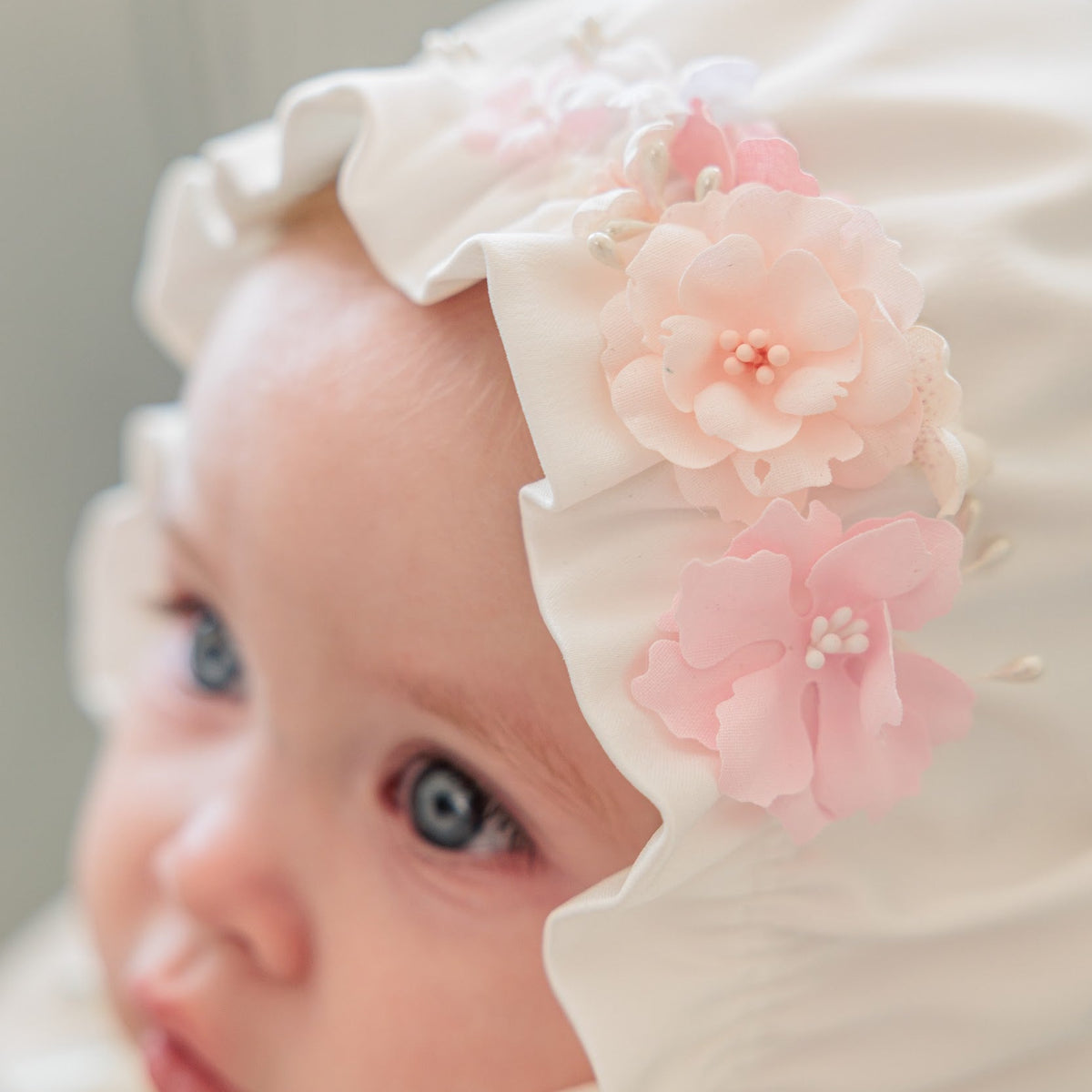 A close-up of a baby wearing a white bonnet with ruffled edges and large pink fabric flowers with small pearl-like centers.