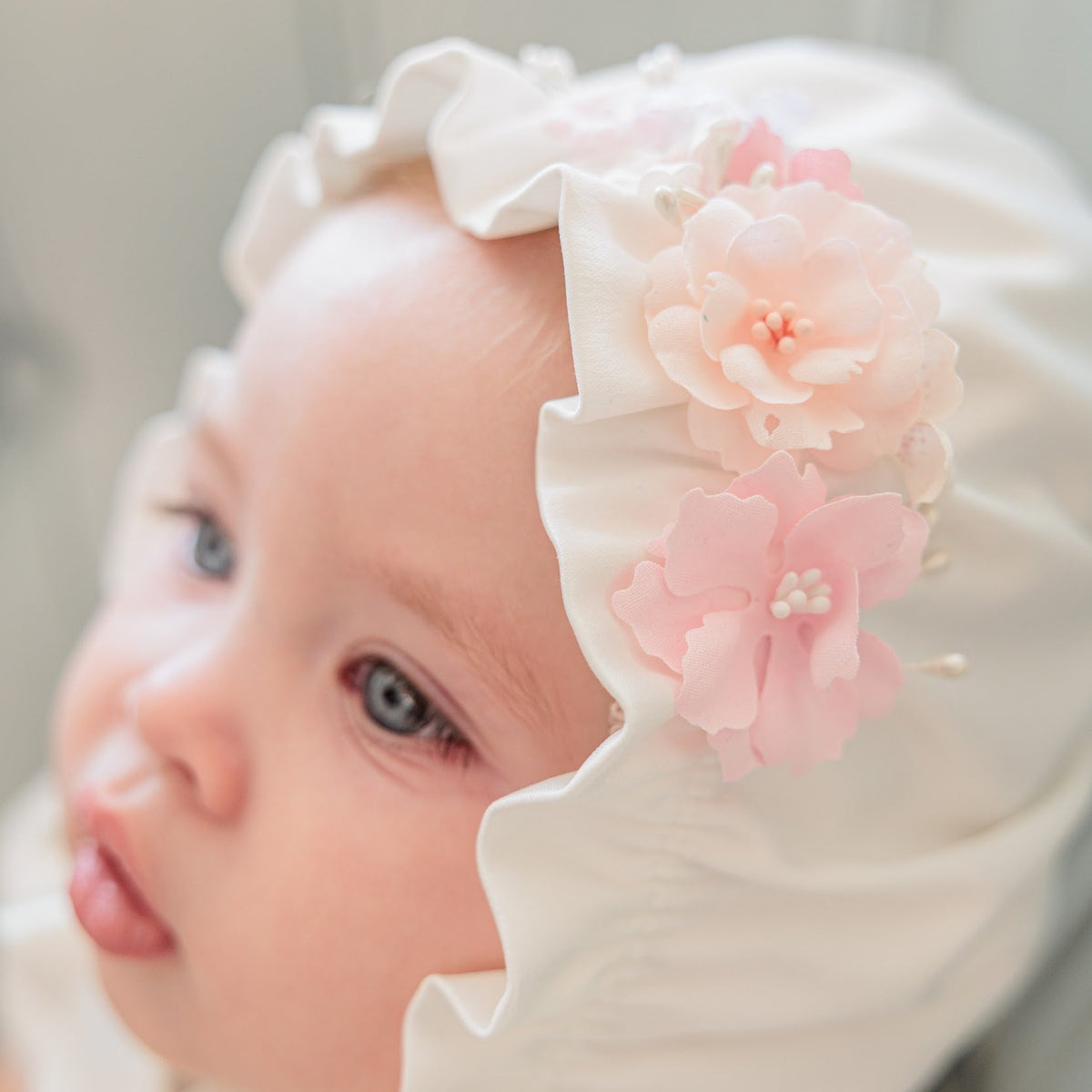 A close-up of a baby wearing a cream-colored bonnet with two soft pink fabric flowers on the side, set against a soft, neutral background.