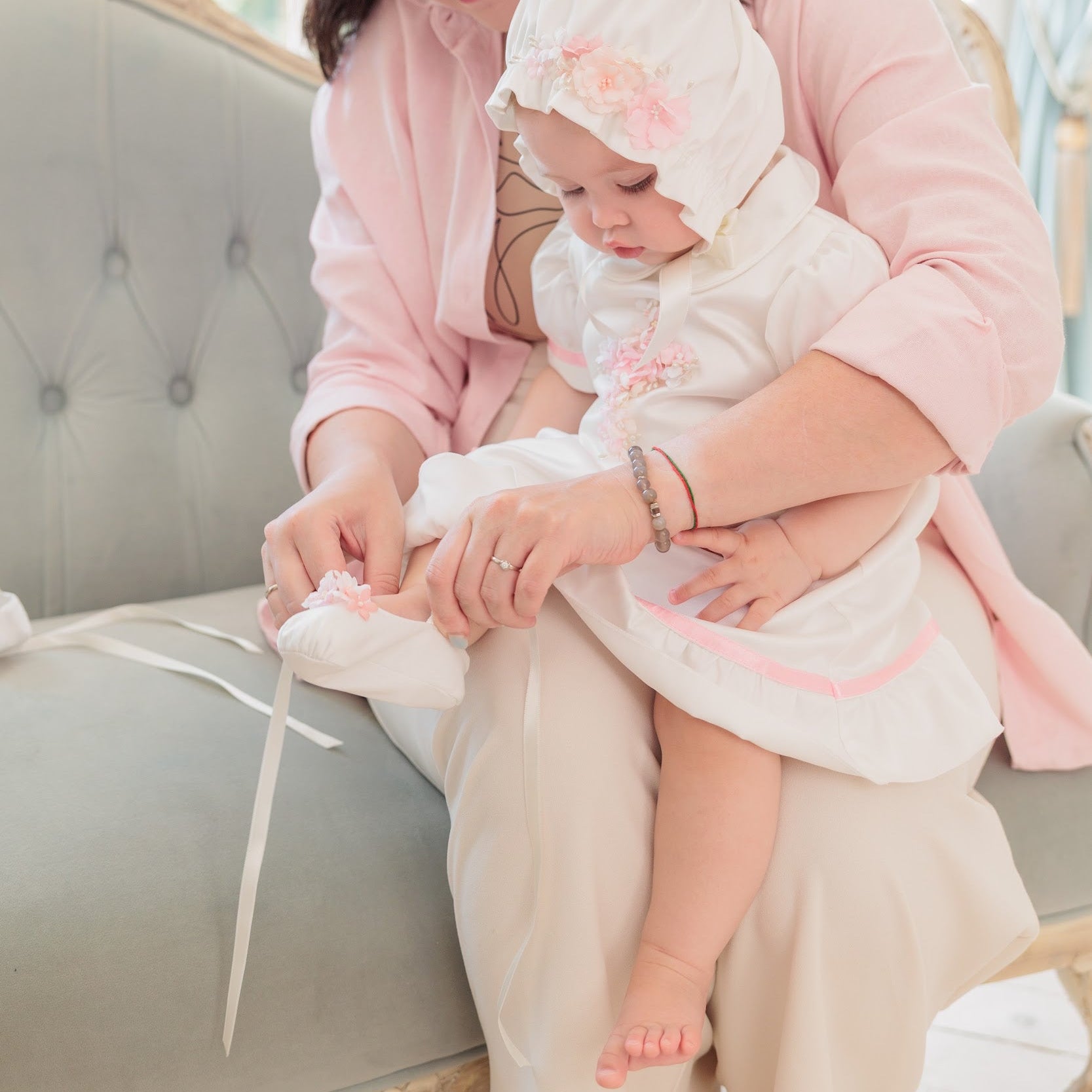 A woman in a pink robe and beige pants sits on a tufted sofa, helping a baby in a white bonnet and white dress with pink trim wear a white bootie.