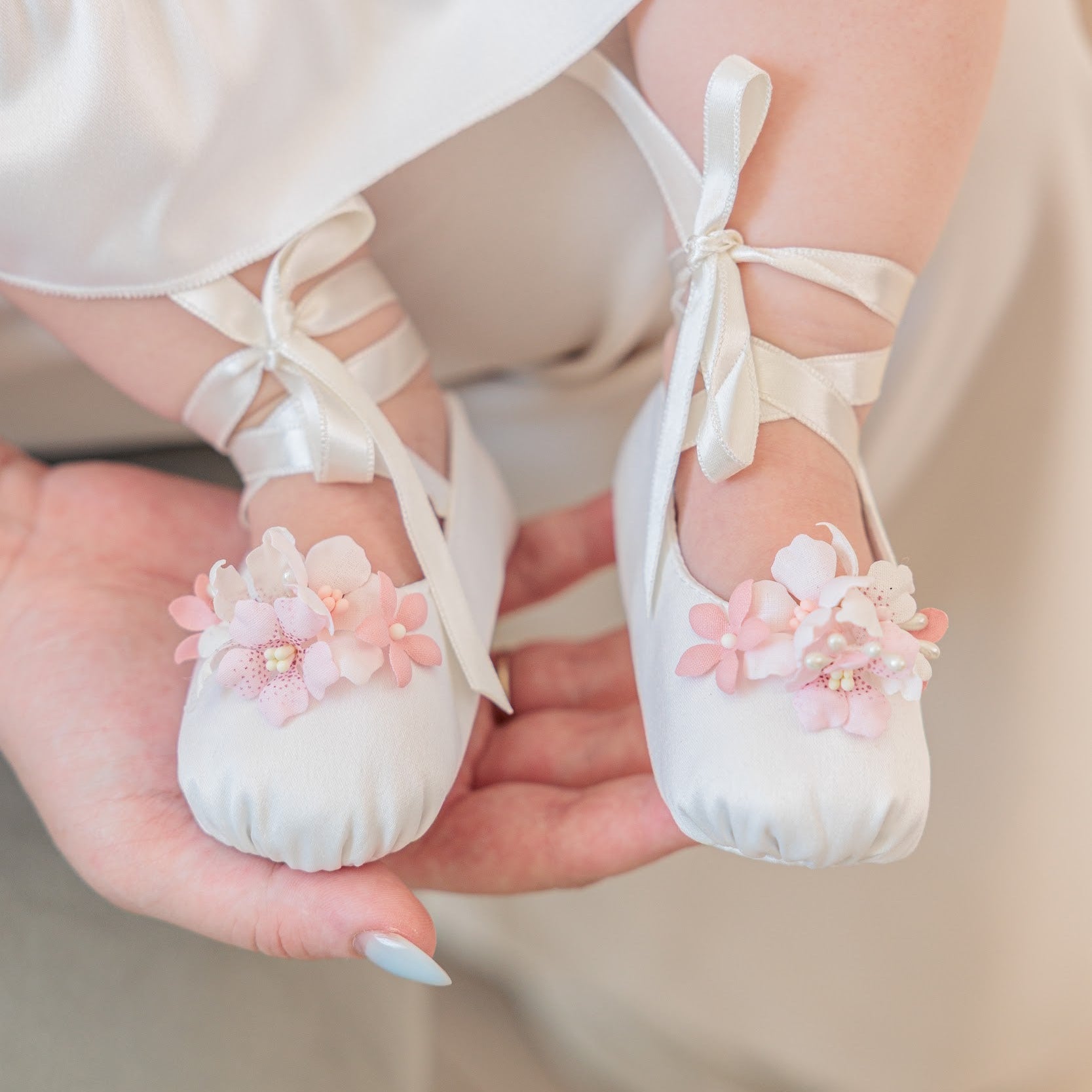 White satin ballet-style baby shoes with pink floral embellishments and ribbon ties, held in a person's hands. The baby is wearing a white dress with a pink waistband.