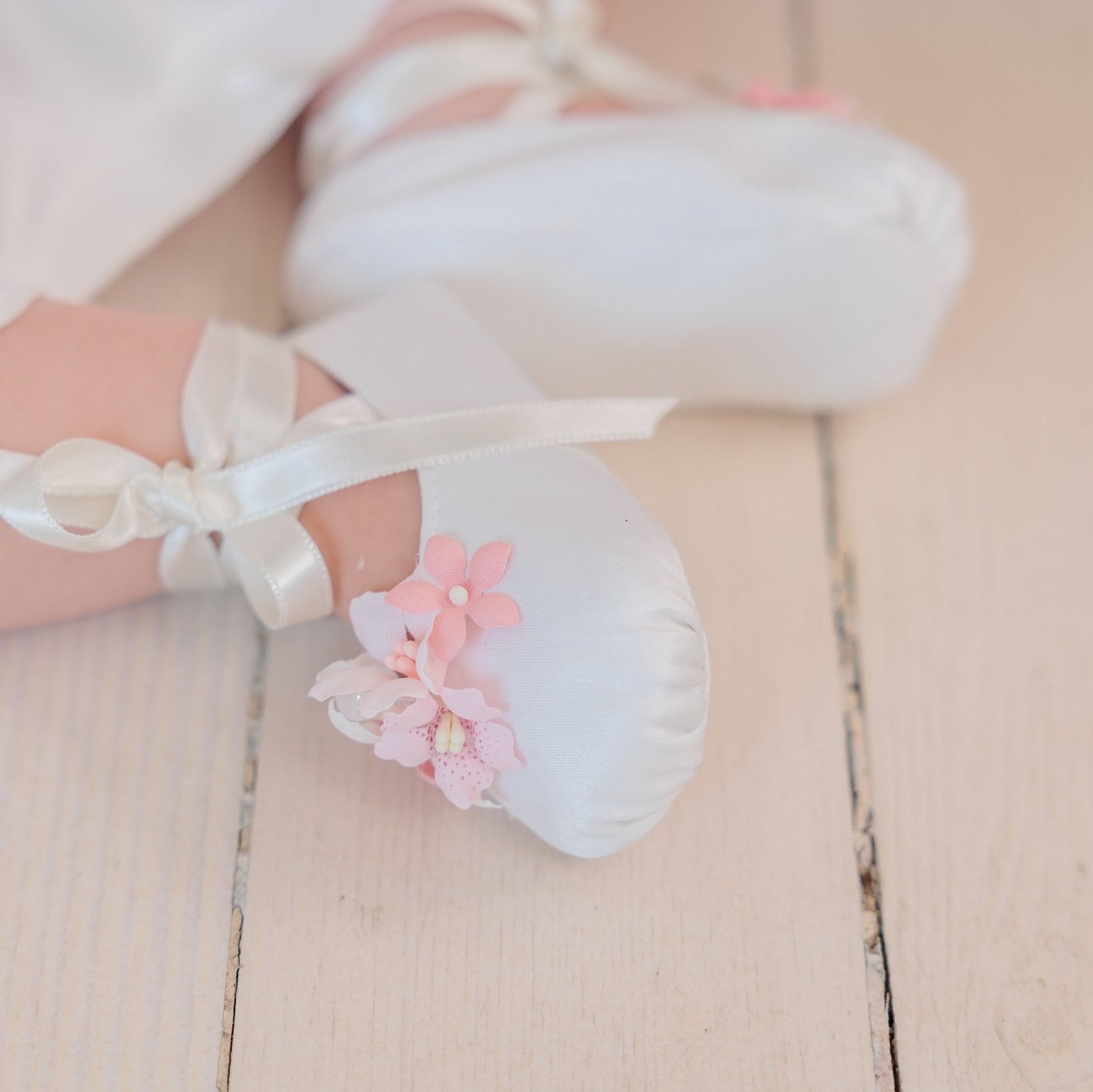 White satin ballet slippers with white ribbon ties and pink fabric flowers, shown on a child's feet on a light wooden floor.