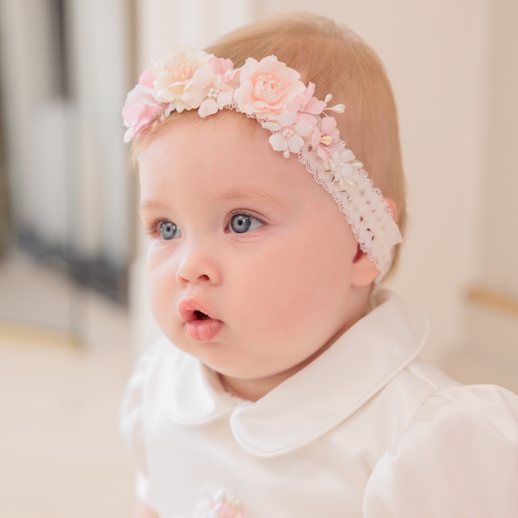 A baby girl with light hair and blue eyes wearing a pink and white floral headband with a lace trim, paired with a white short-sleeve dress with a Peter Pan collar.