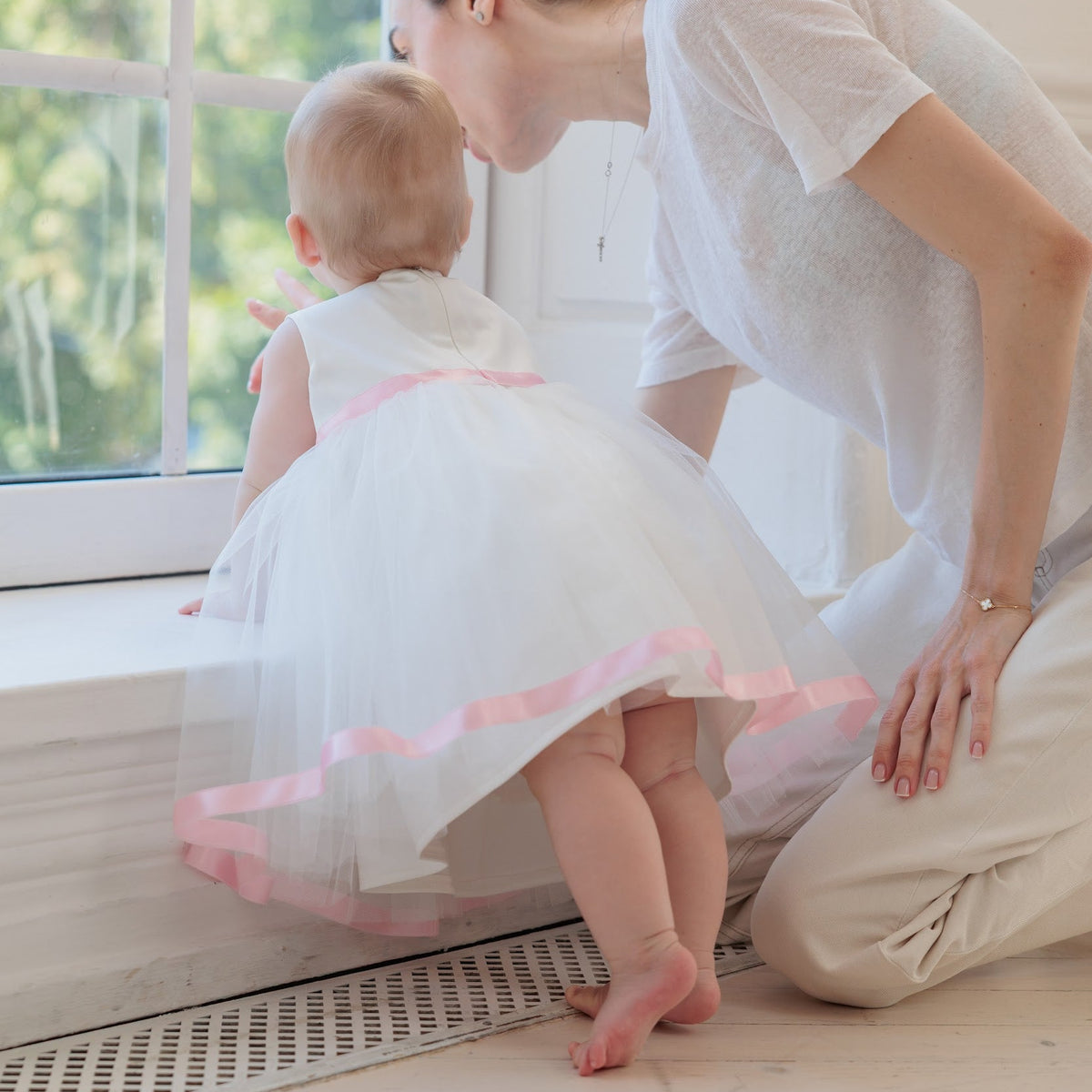 A woman kneels beside a baby in a white tutu dress with pink trim, looking at the child by a bright window in a light, airy room.
