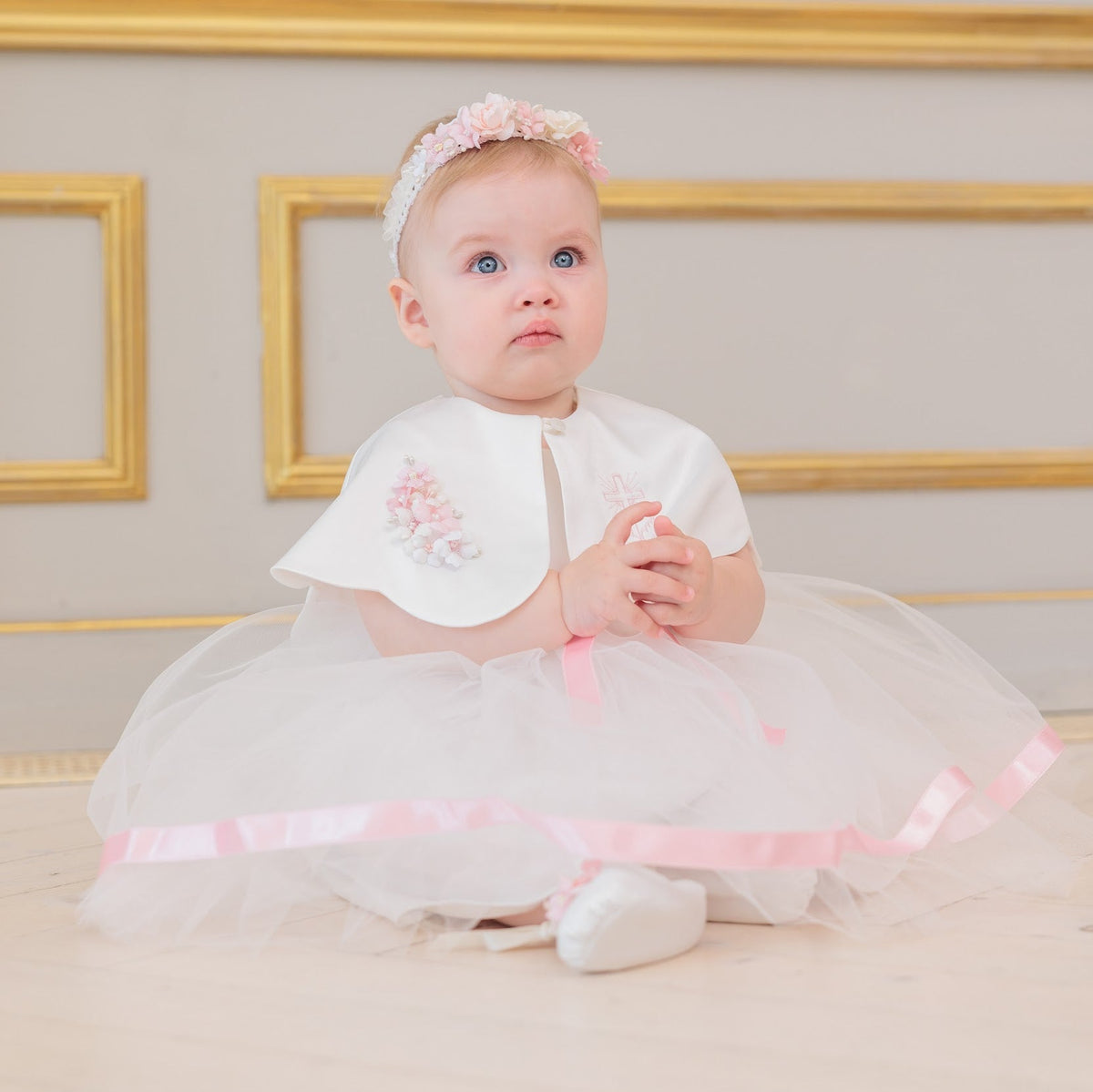 A baby girl wearing a white and pink first birthday tutu dress with a matching headband, sitting on a light wooden floor in front of a beige wall with gold picture frames.