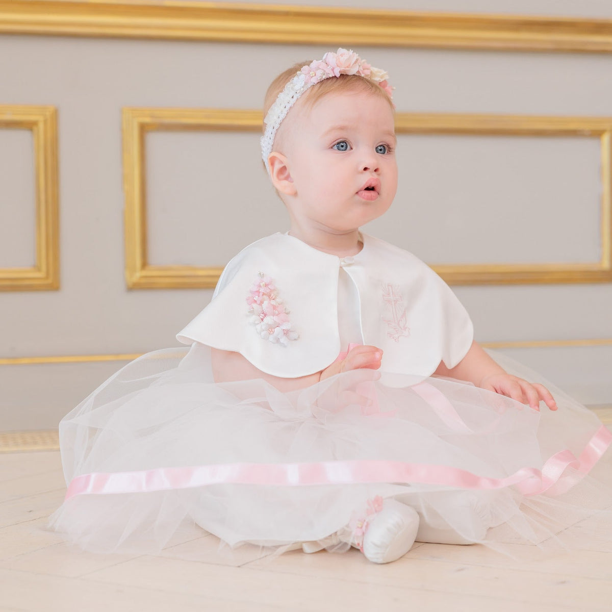 A baby girl wearing a white and pink first birthday dress with a tulle skirt, a white collar, and a matching pink headband, sitting on a light wooden floor in front of a beige wall with gold picture frames.